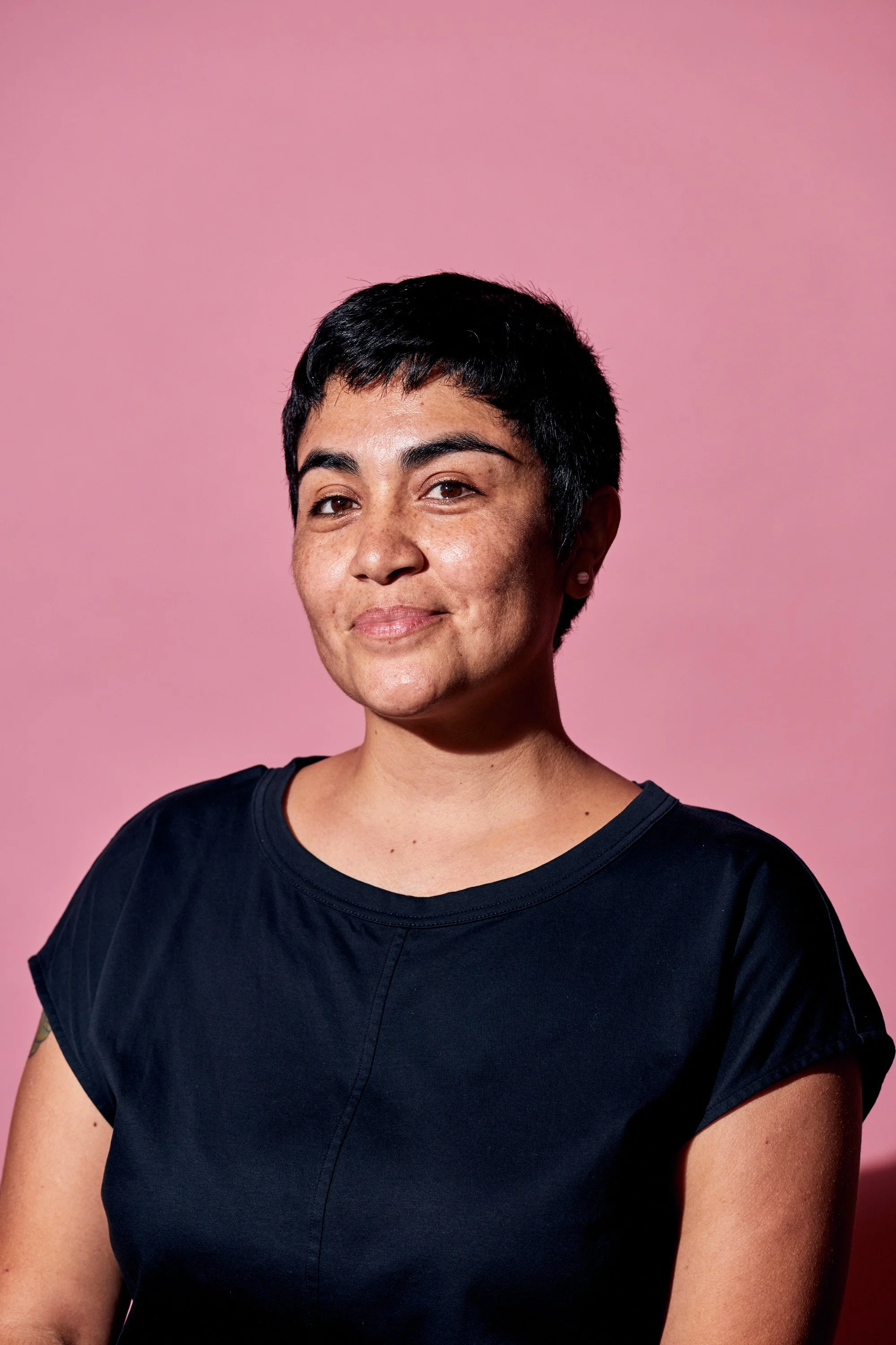 A woman with short black hair, wearing a black top, stands against a pink background, smiling gently.