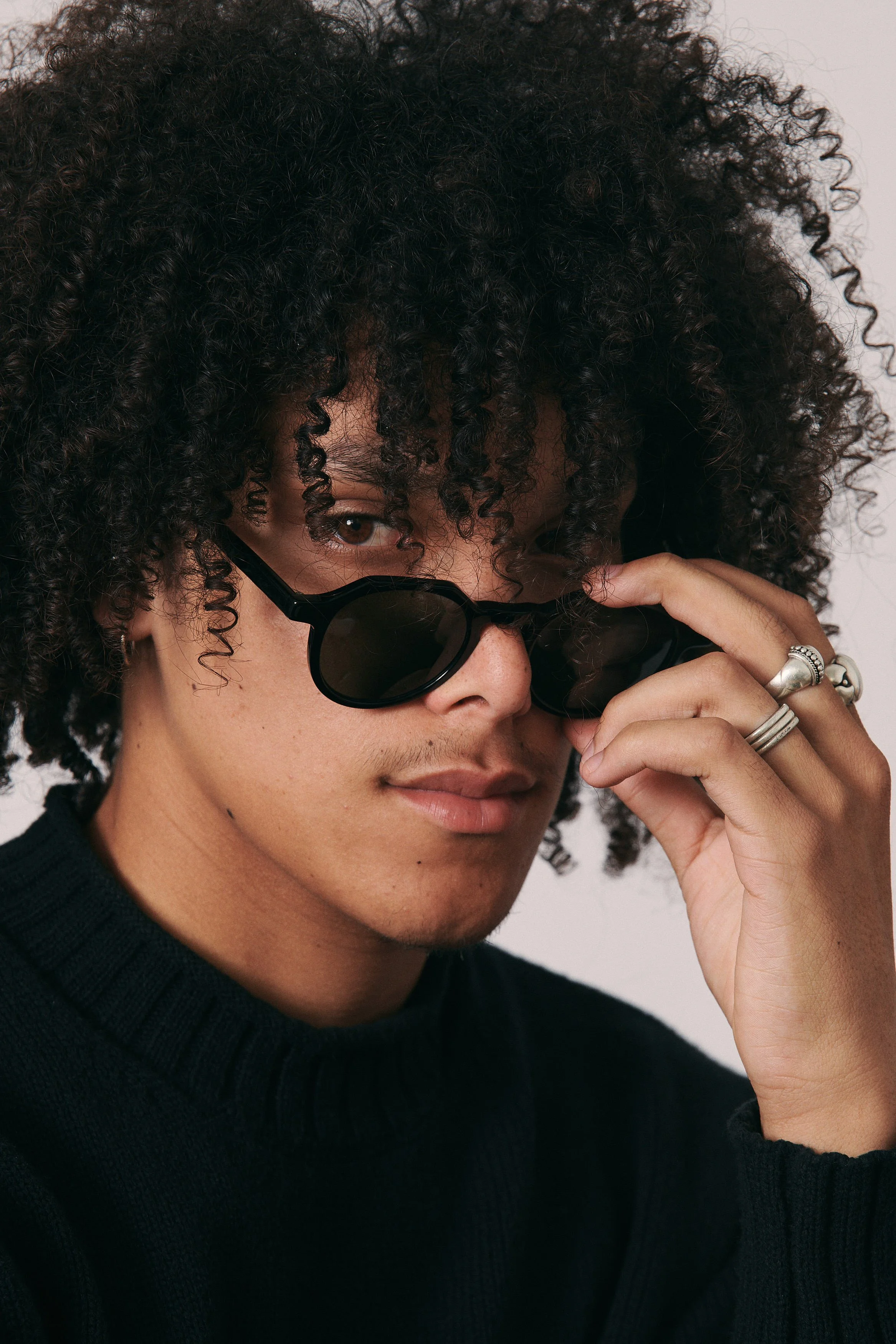 A person with curly hair wearing black sunglasses and a black sweater, adjusting their sunglasses with their right hand, which has multiple silver rings.