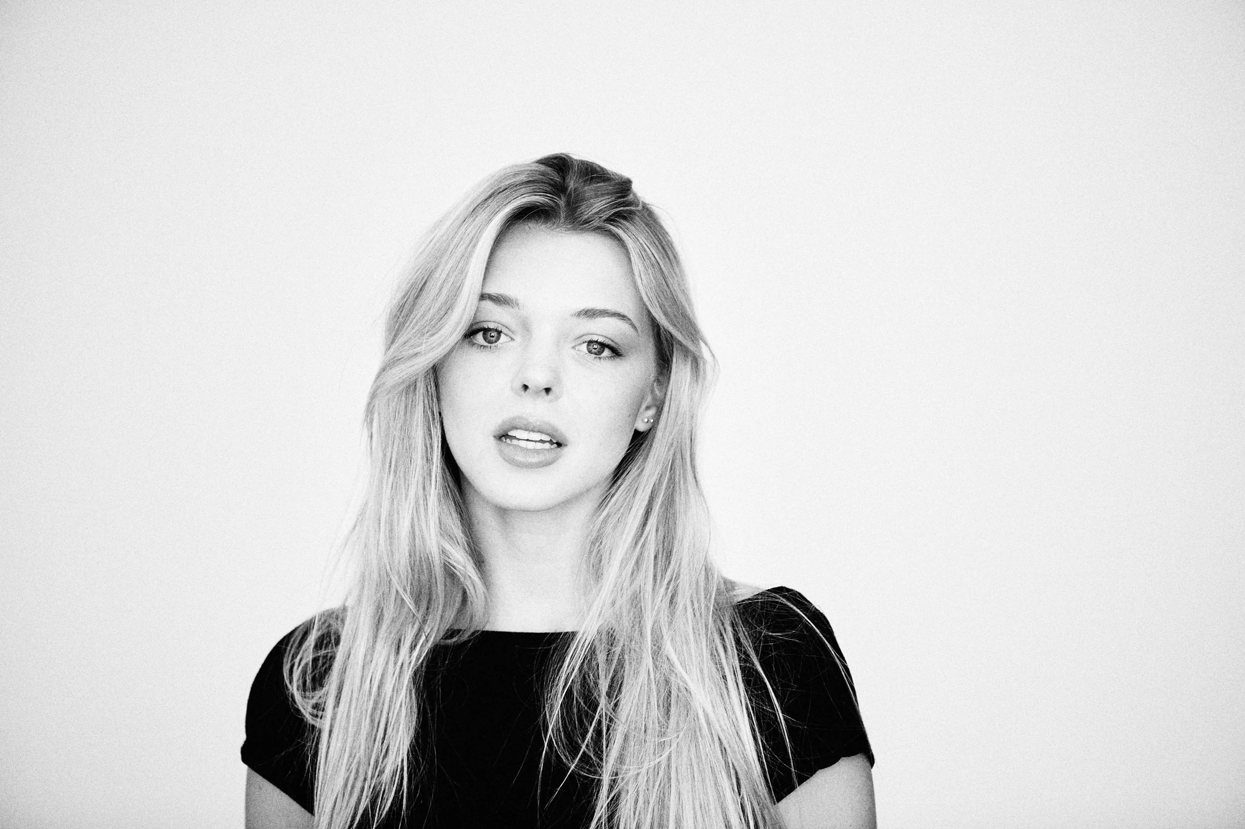 Black and white portrait of a woman with long wavy hair, wearing a black top, looking directly at the camera with a neutral expression.