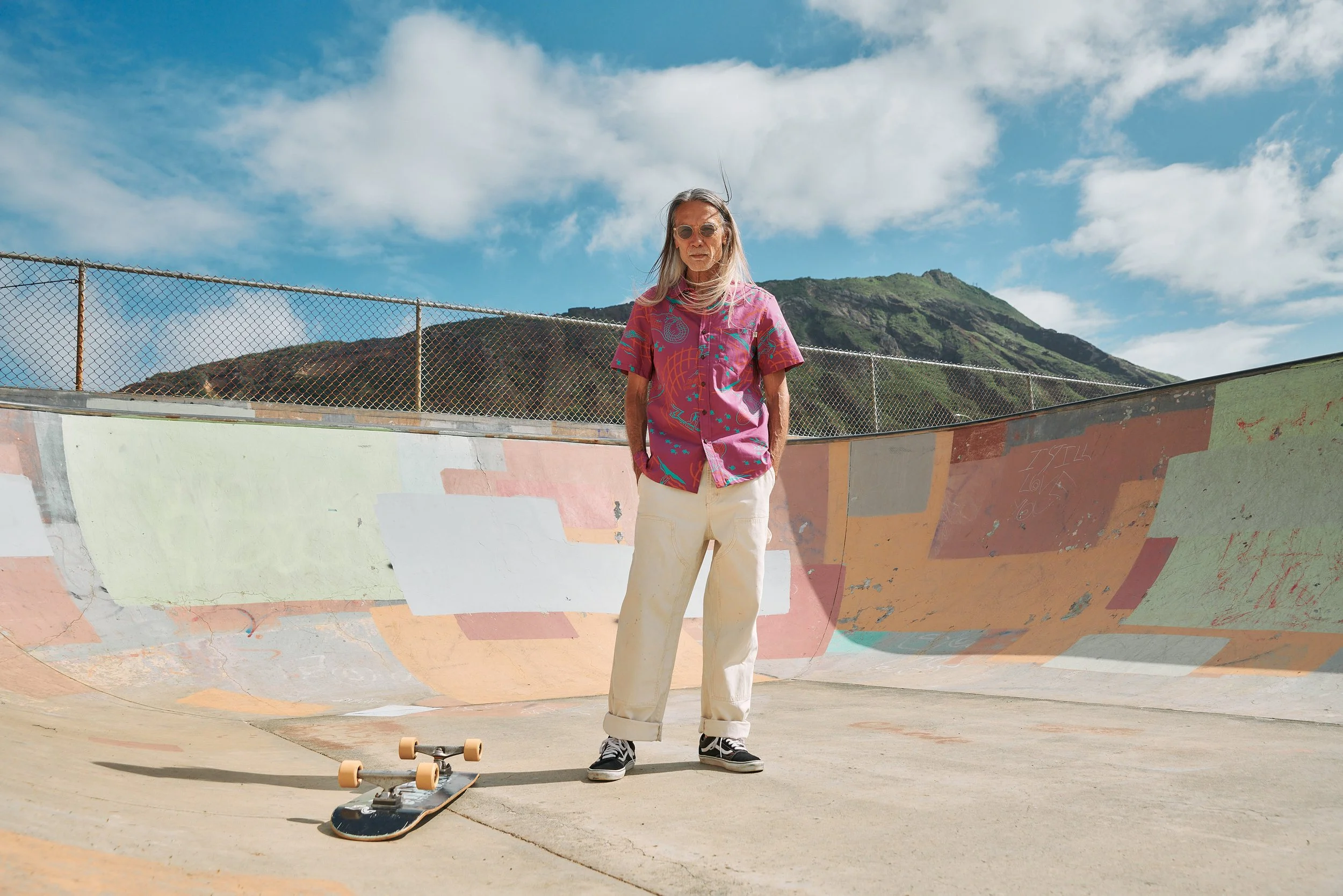 An elderly woman with long gray hair, wearing sunglasses, a vibrant pink patterned shirt, beige pants, and black and white sneakers, stands with her hands in her pockets at a skate park, next to a skateboard lying on the ground. Behind her is a concr