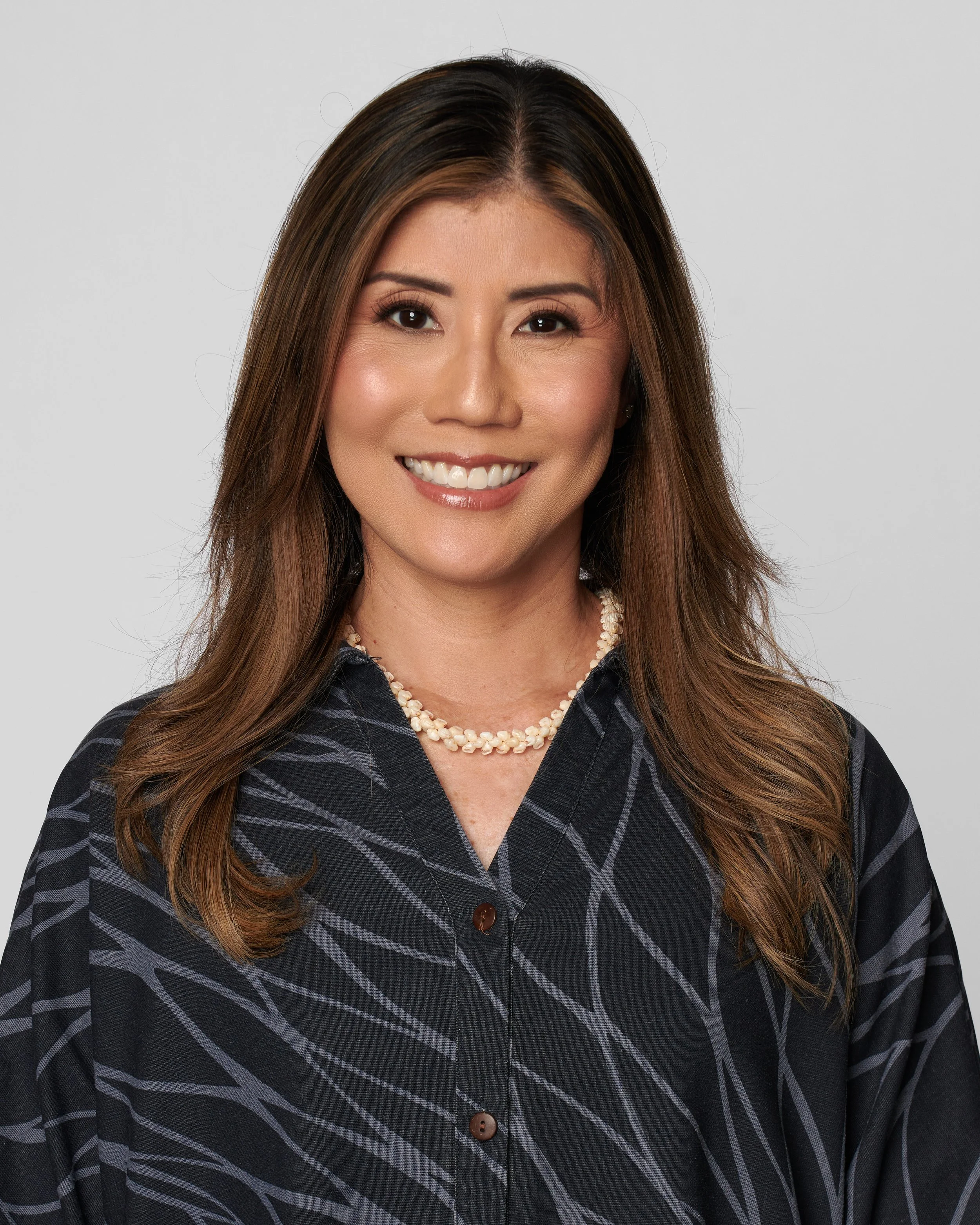 Portrait of an Asian woman with long, wavy brown hair, smiling, wearing a dark patterned blouse and a pearl necklace.