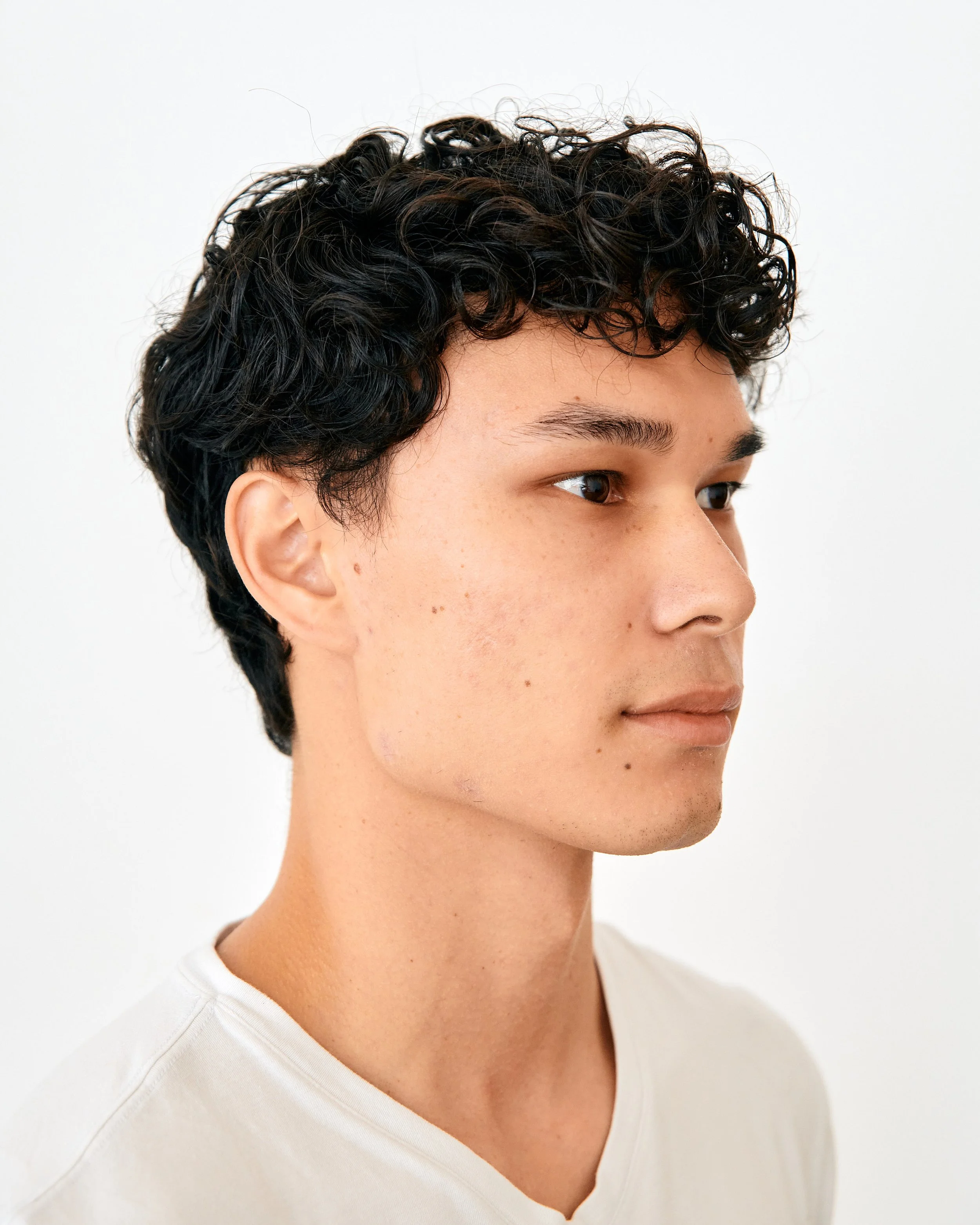 Side profile of a young person with short, curly black hair and light skin, wearing a white shirt.