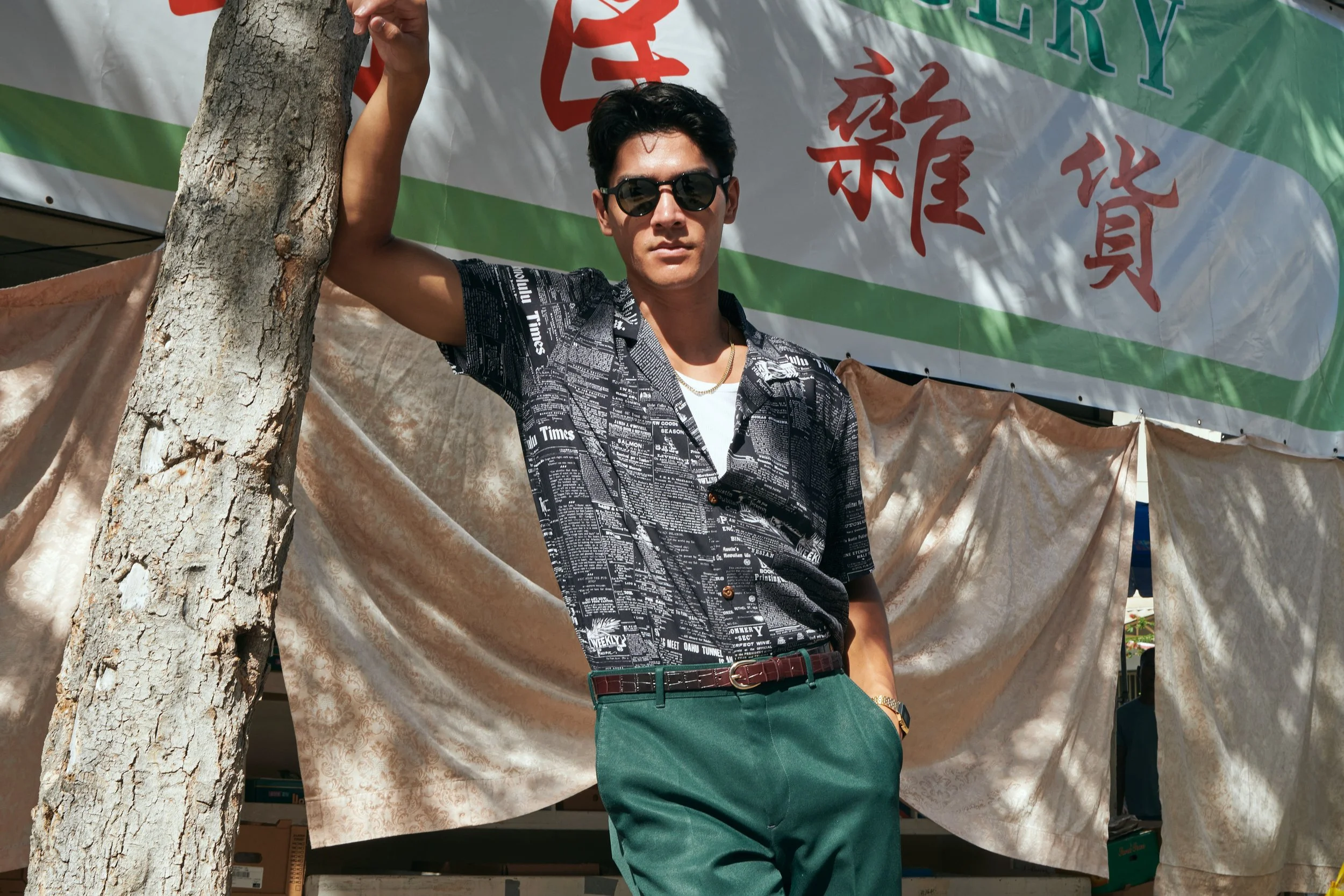 A young man wearing sunglasses, a black patterned short-sleeve shirt, green pants, and a white t-shirt underneath, standing outdoors behind a tree, with a banner and fabric backdrop in the background.