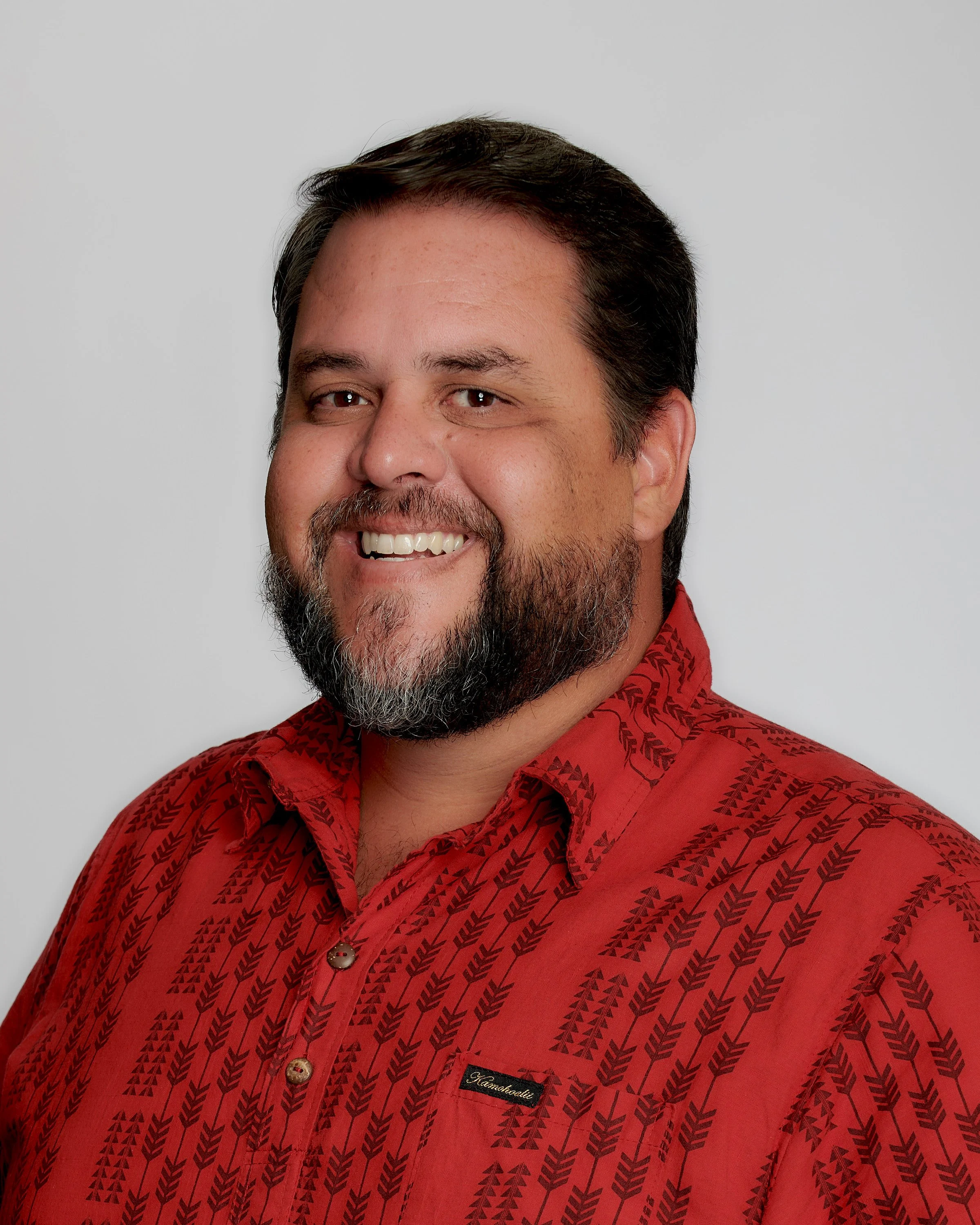 Portrait of a smiling man with dark hair, a beard, and mustache wearing a red patterned shirt against a plain gray background.