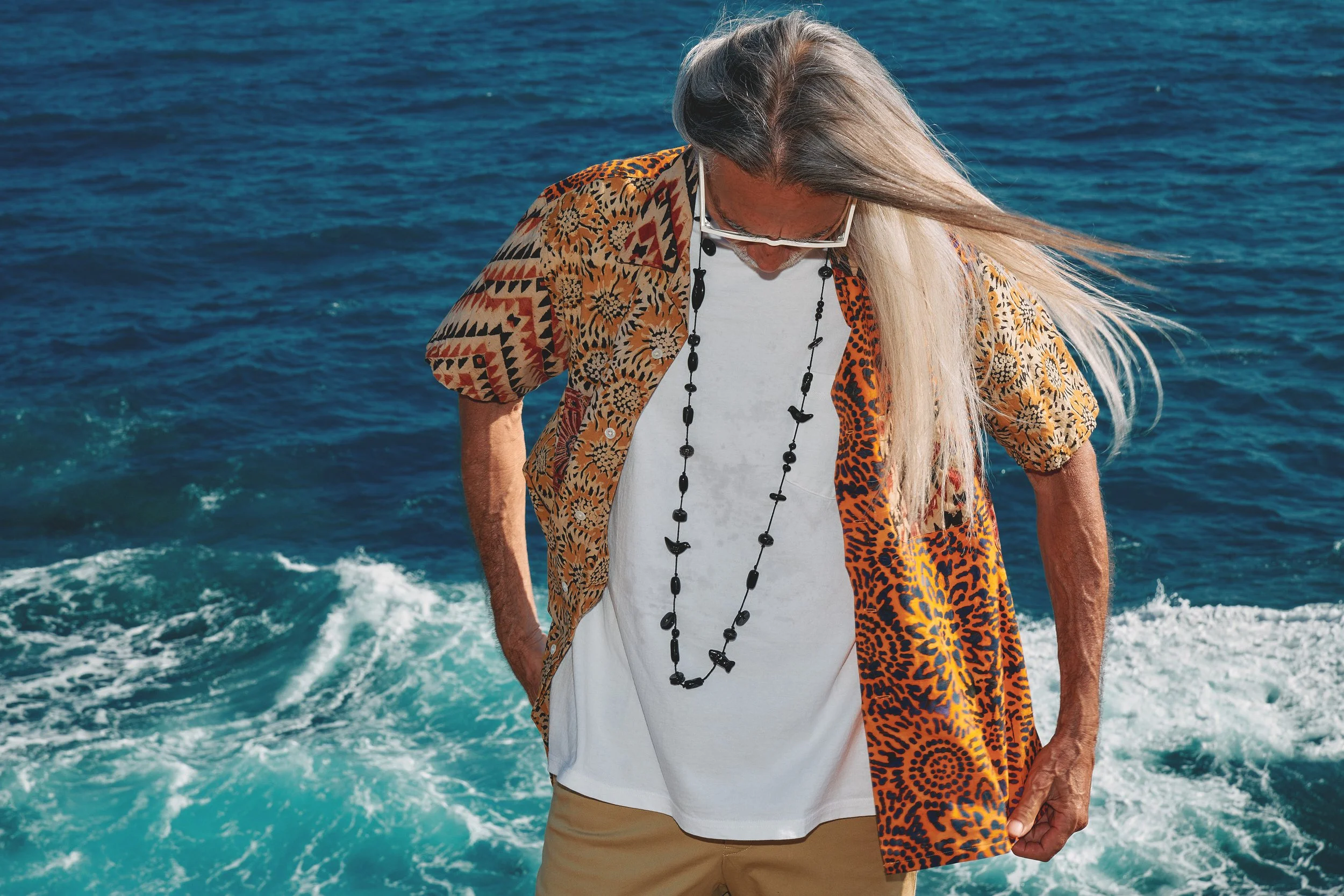 An elderly man with long, gray hair standing on a boat with the ocean in the background, wearing sunglasses, a colorful animal print shirt, a white T-shirt, khaki pants, and a black beaded necklace.