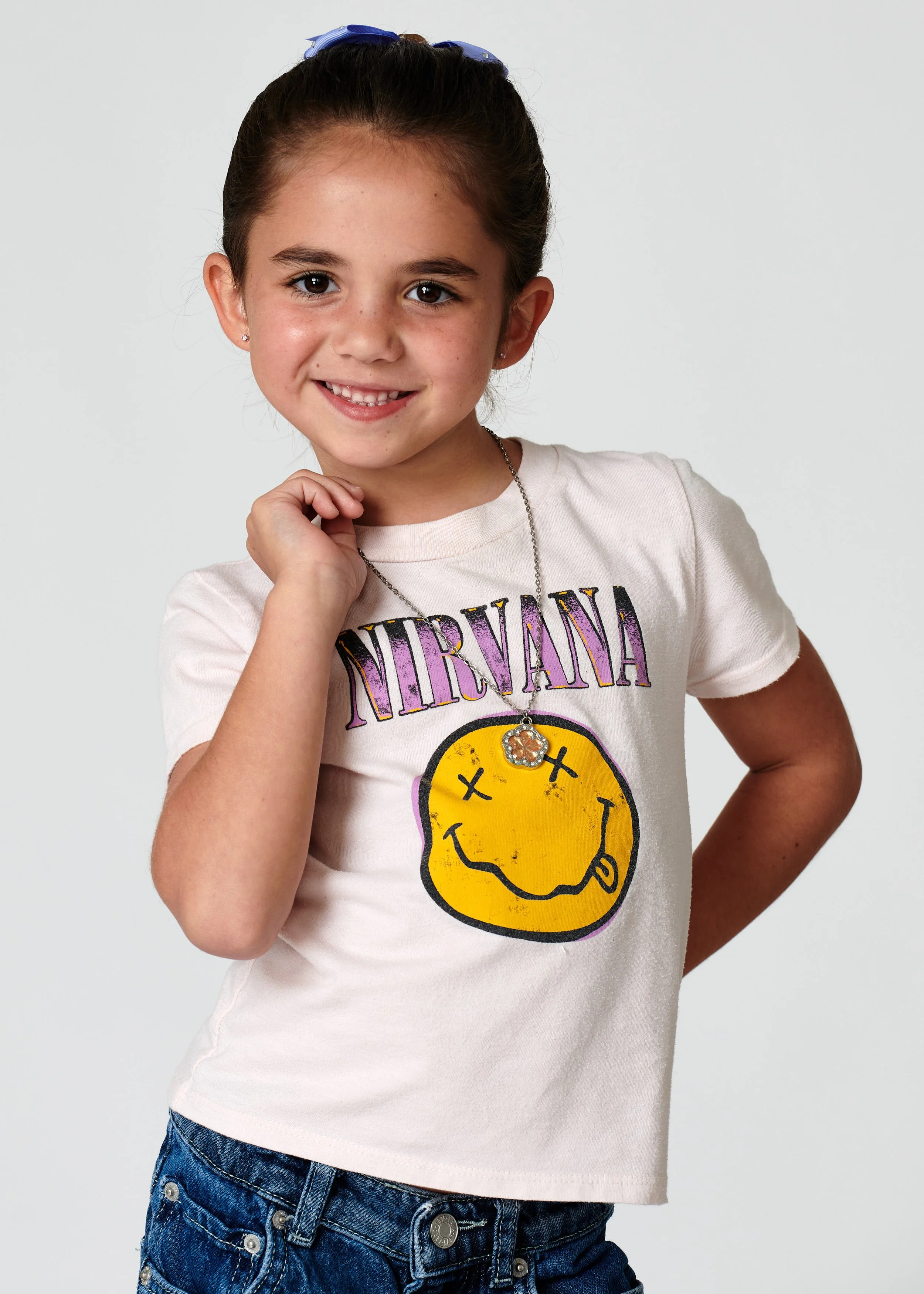 Young girl with brown hair in a ponytail, wearing a Nirvana t-shirt and denim shorts, smiling and posing with her right hand near her face on a plain light background.