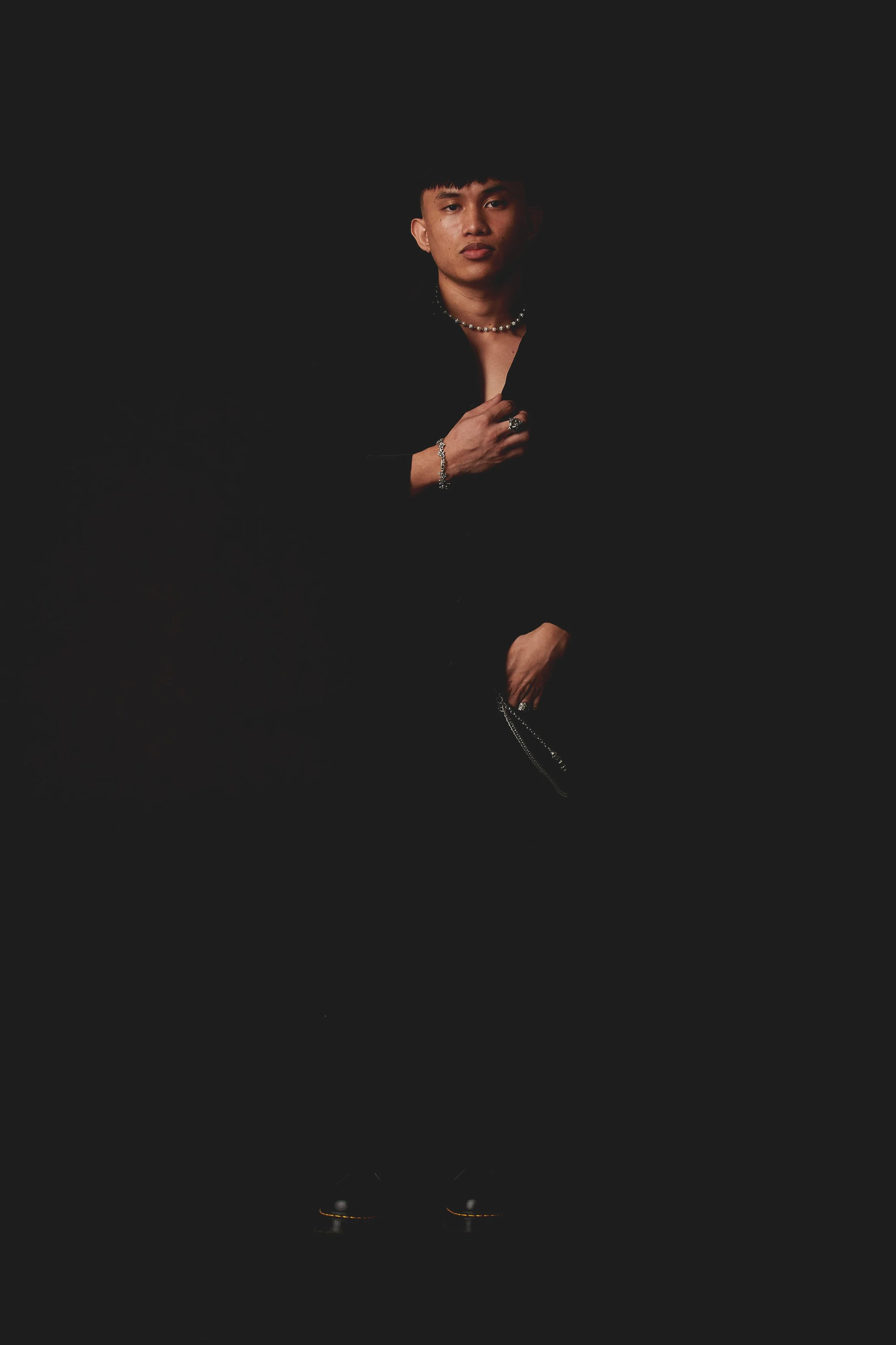 A person dressed in black with jewelry, standing against a dark background.