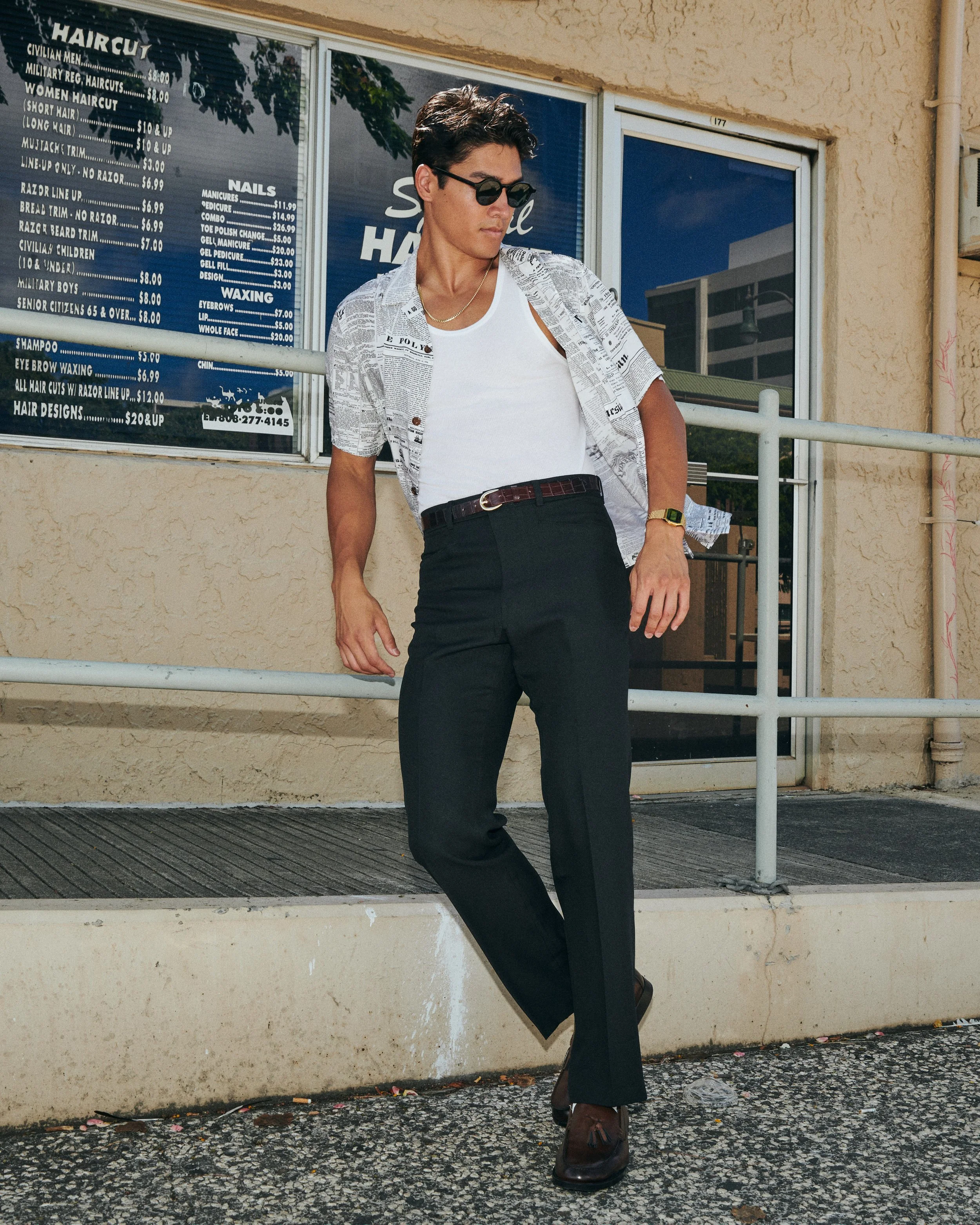 A person with short dark hair, wearing sunglasses, a white tank top, black pants, and brown shoes, standing outside a storefront with a menu on the window.