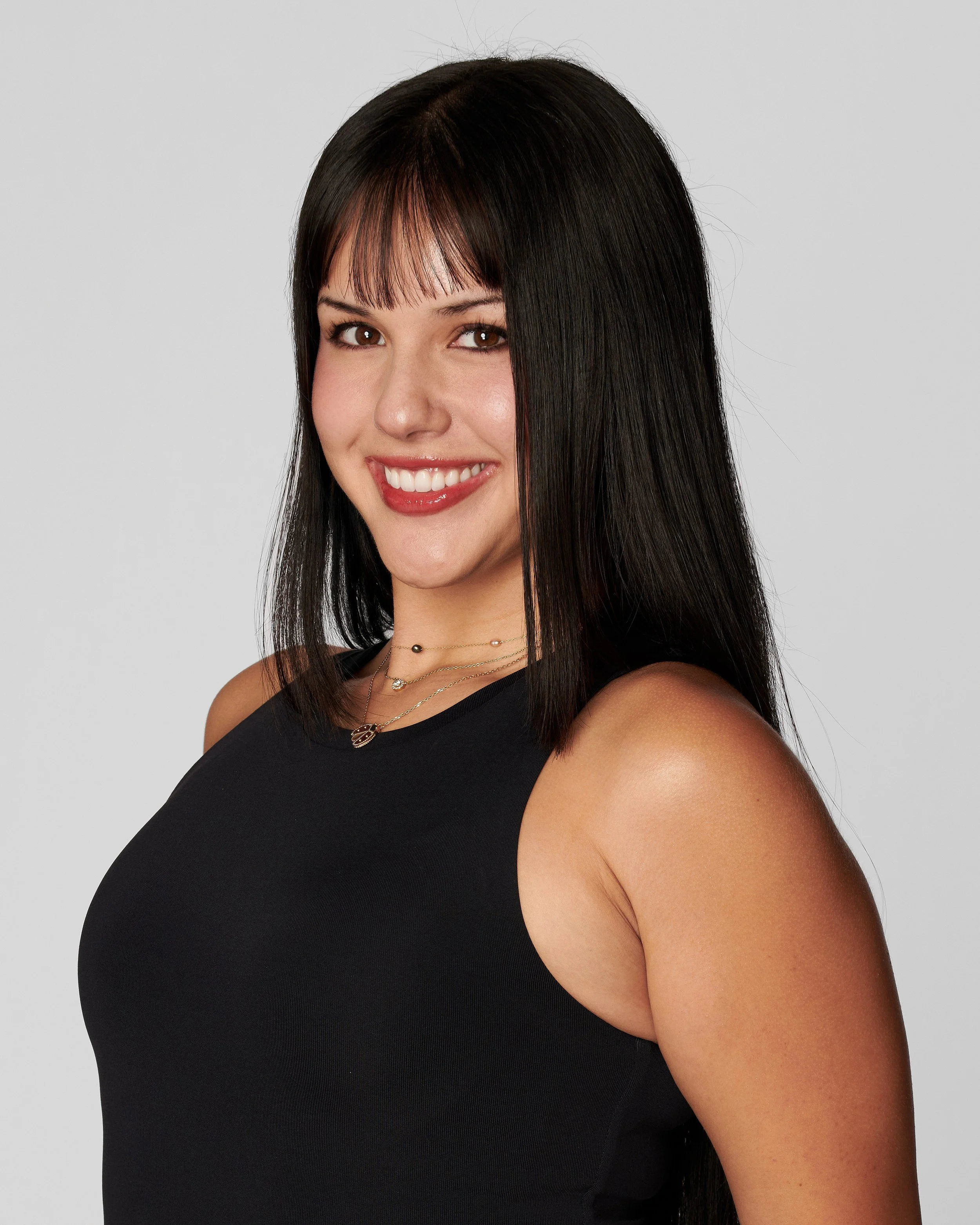 A young woman with long black hair, smiling, wearing a black sleeveless top and layered necklaces, against a plain gray background.