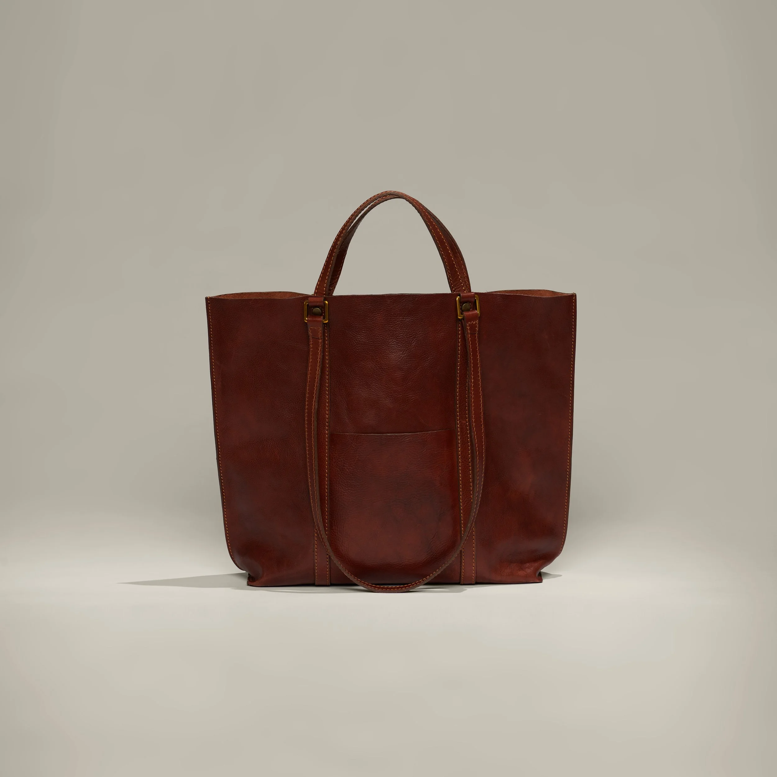 A large, brown leather tote bag with double handles, featuring a front pocket, set against a plain, light background.