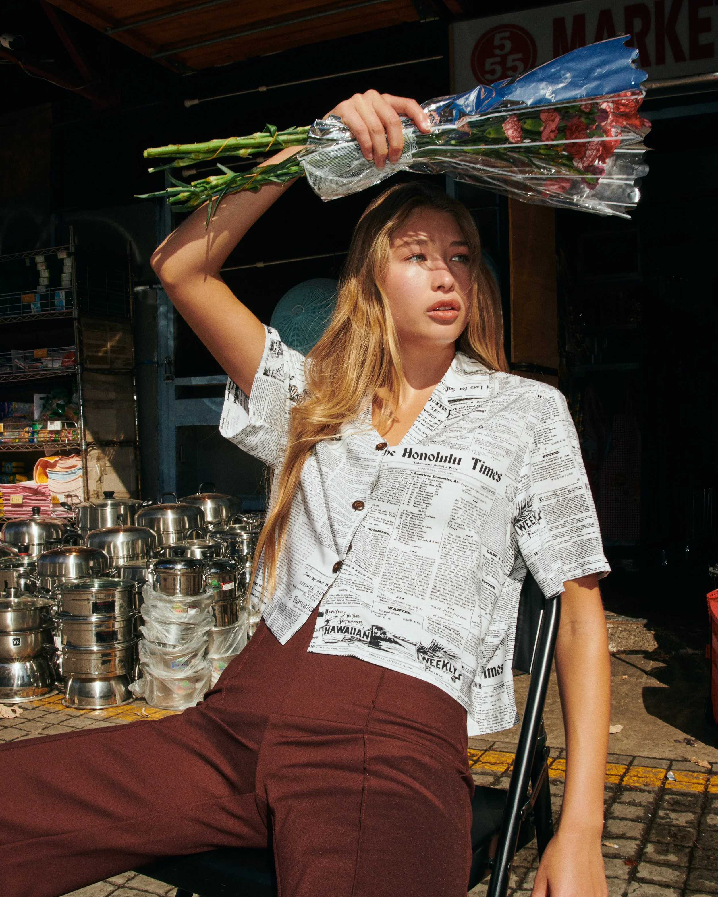 A woman sitting outdoors at a market, holding a bouquet of red flowers above her head. She has long blonde hair and is wearing a white shirt with newspaper print and maroon pants.