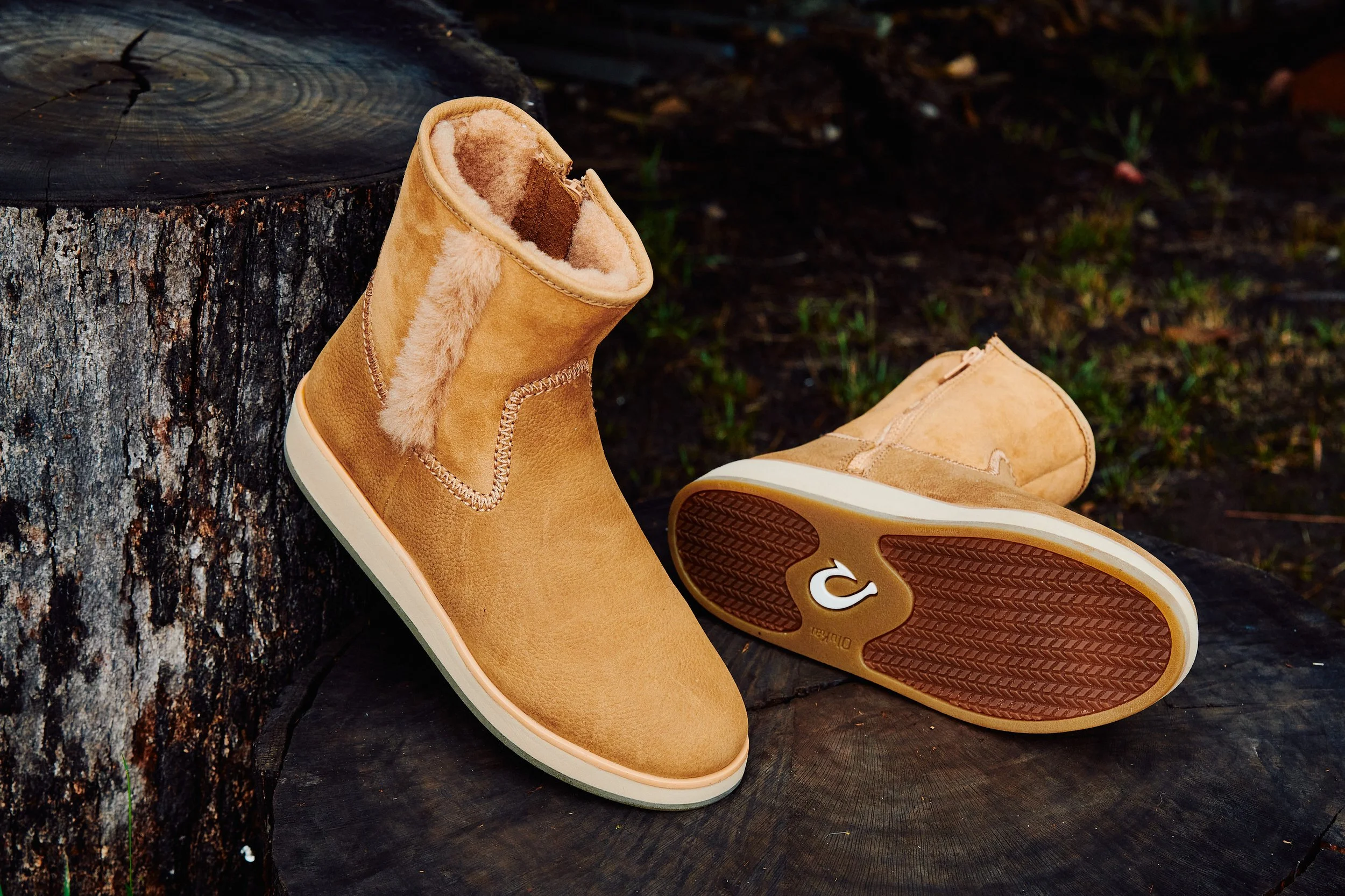 Tan shearling-lined boots placed on a tree stump outdoors, with one upright and the other lying on its side showing the sole with a logo.