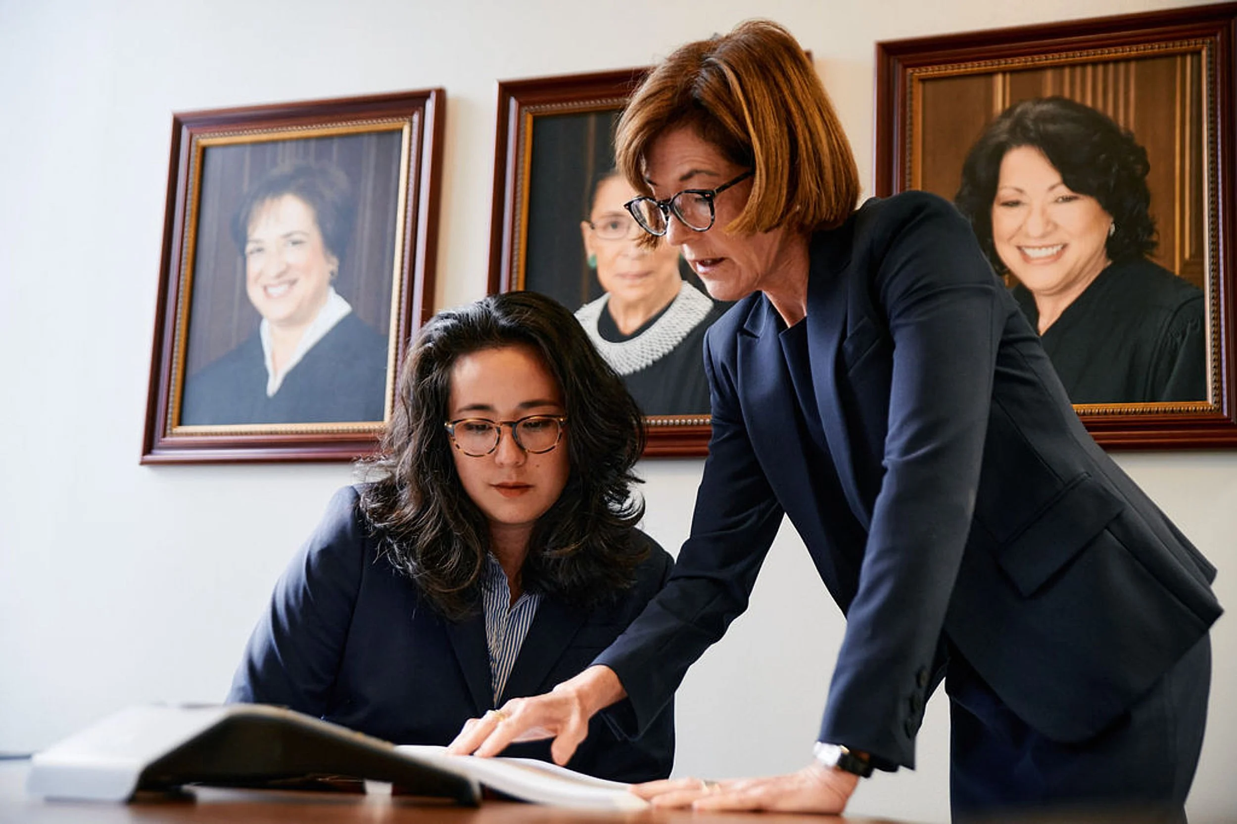 A woman with glasses in a navy blazer and a young woman with glasses seated at a desk looking at documents, with an older woman with glasses standing and pointing at the papers. Portraits of women in legal attire hang on the wall behind them.