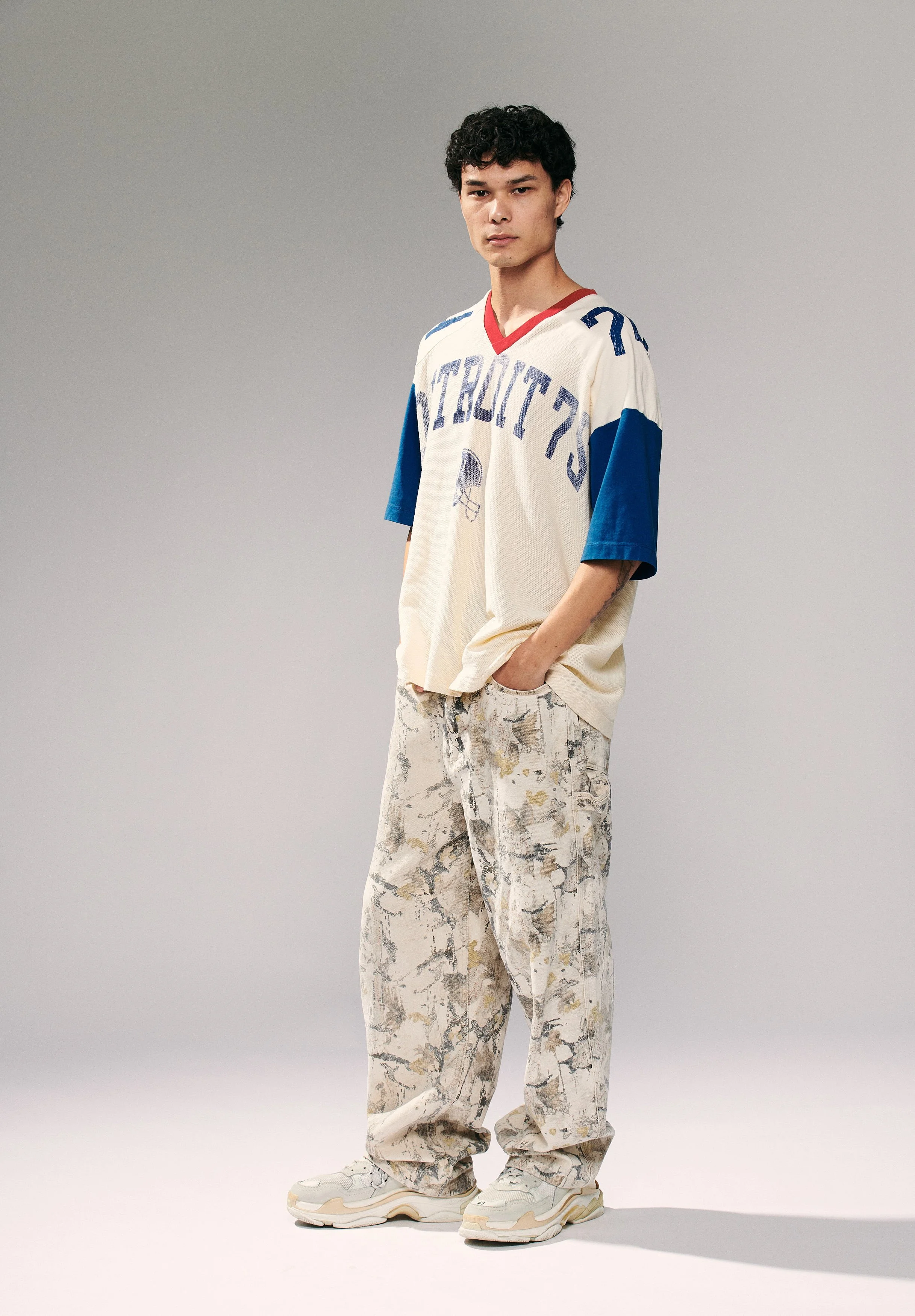 Young man with curly black hair in a vintage Detroit sports jersey, patterned baggy pants, and sneakers, standing against a plain wall.