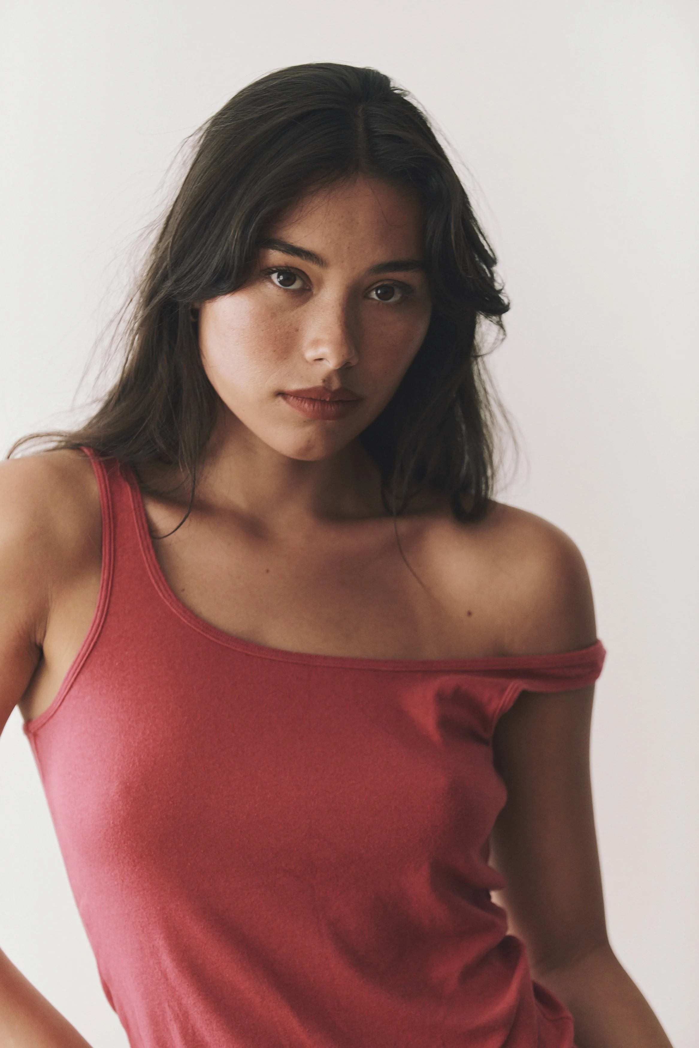 Portrait of a young woman with dark brown hair, wearing a red off-the-shoulder tank top, looking directly at the camera against a plain light background.