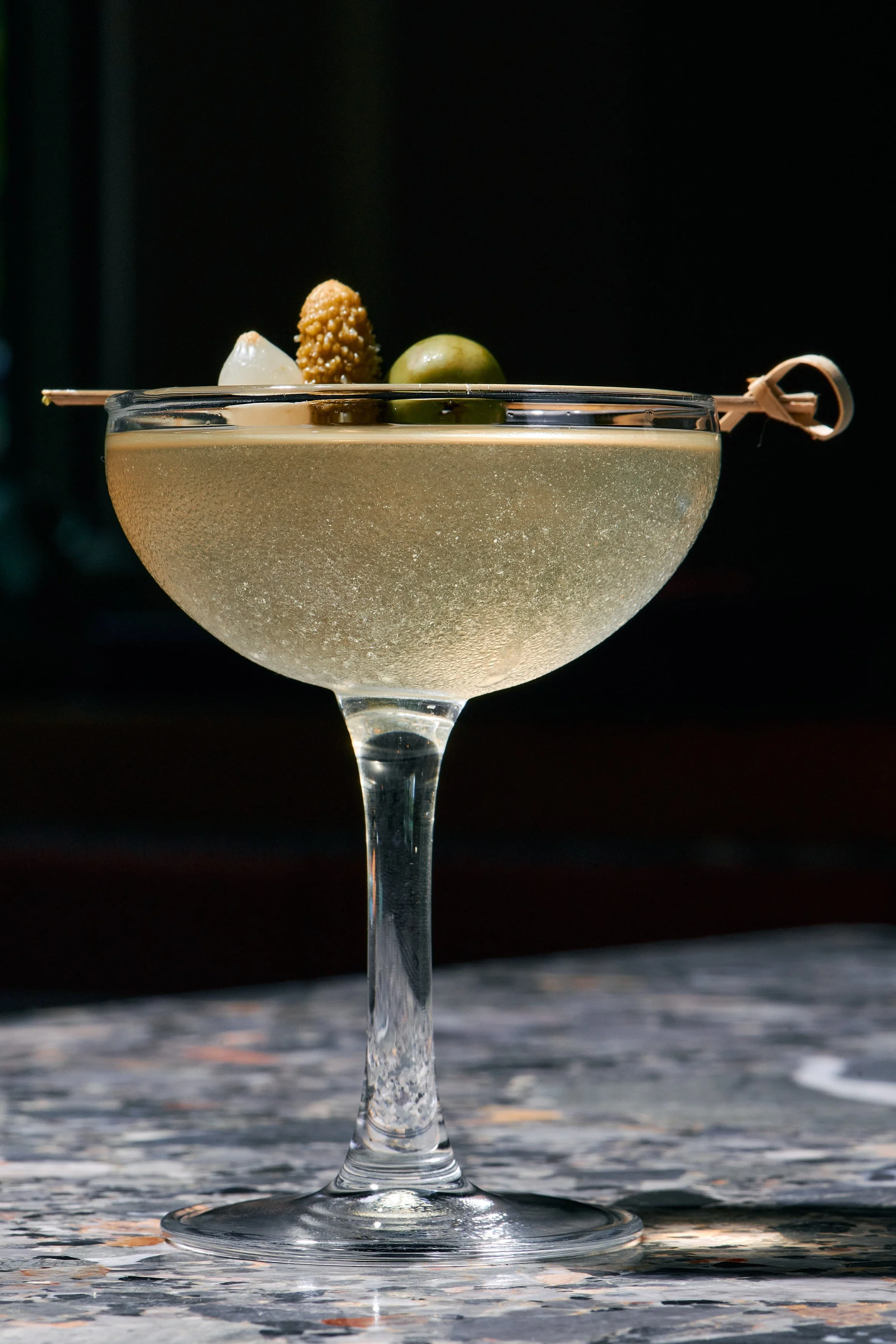 A cocktail in a coupe glass garnished with olives, a pickled onion, and a sesame-coated snack, set on a marble surface against a dark background.