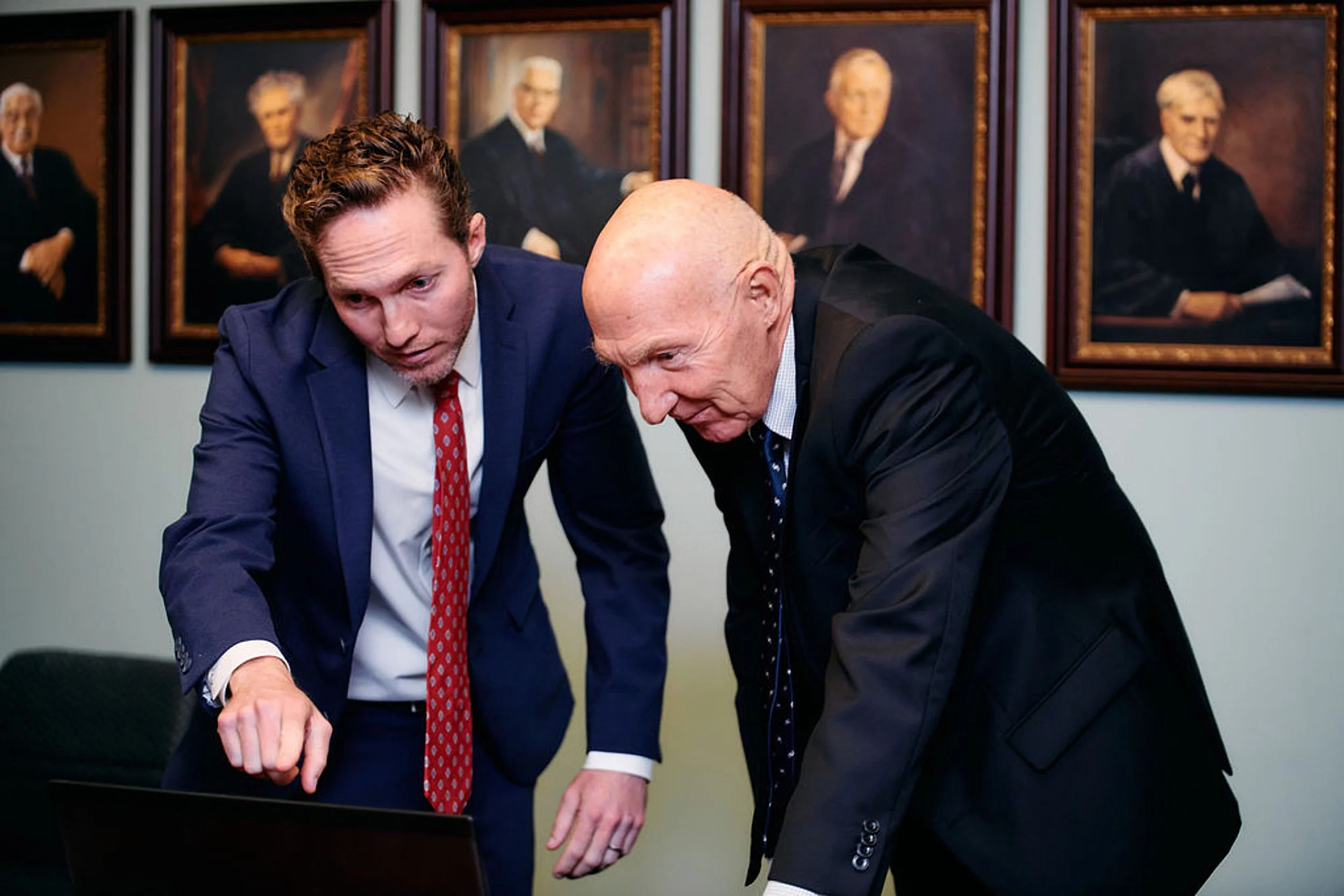 Two men in suits talking and pointing at a laptop in an office with framed portraits of elderly individuals on the wall.