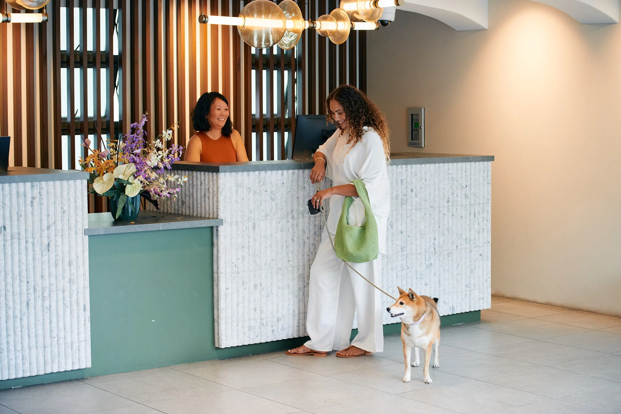 A woman with curly hair and light skin in white pajamas holding a green tote bag and leash with a Shiba Inu dog, checking into a hotel reception with a smiling receptionist behind the counter.