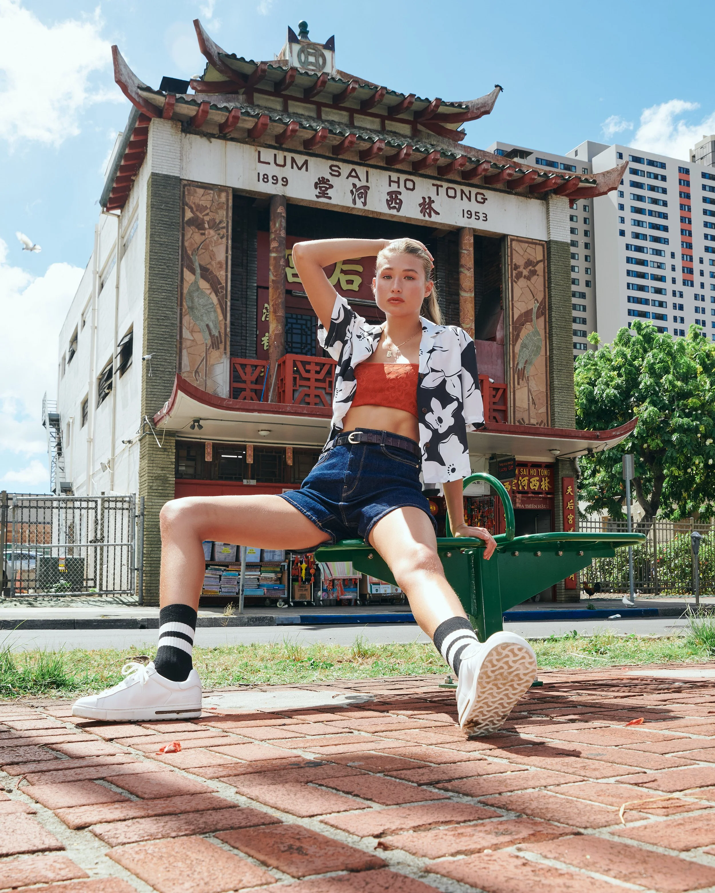 Young woman sitting on a green bench in front of a Chinese-style building with red roof, decorative panels, and Chinese signage, wearing a black and white floral shirt, red top, denim shorts, and white sneakers, under a sunny sky.