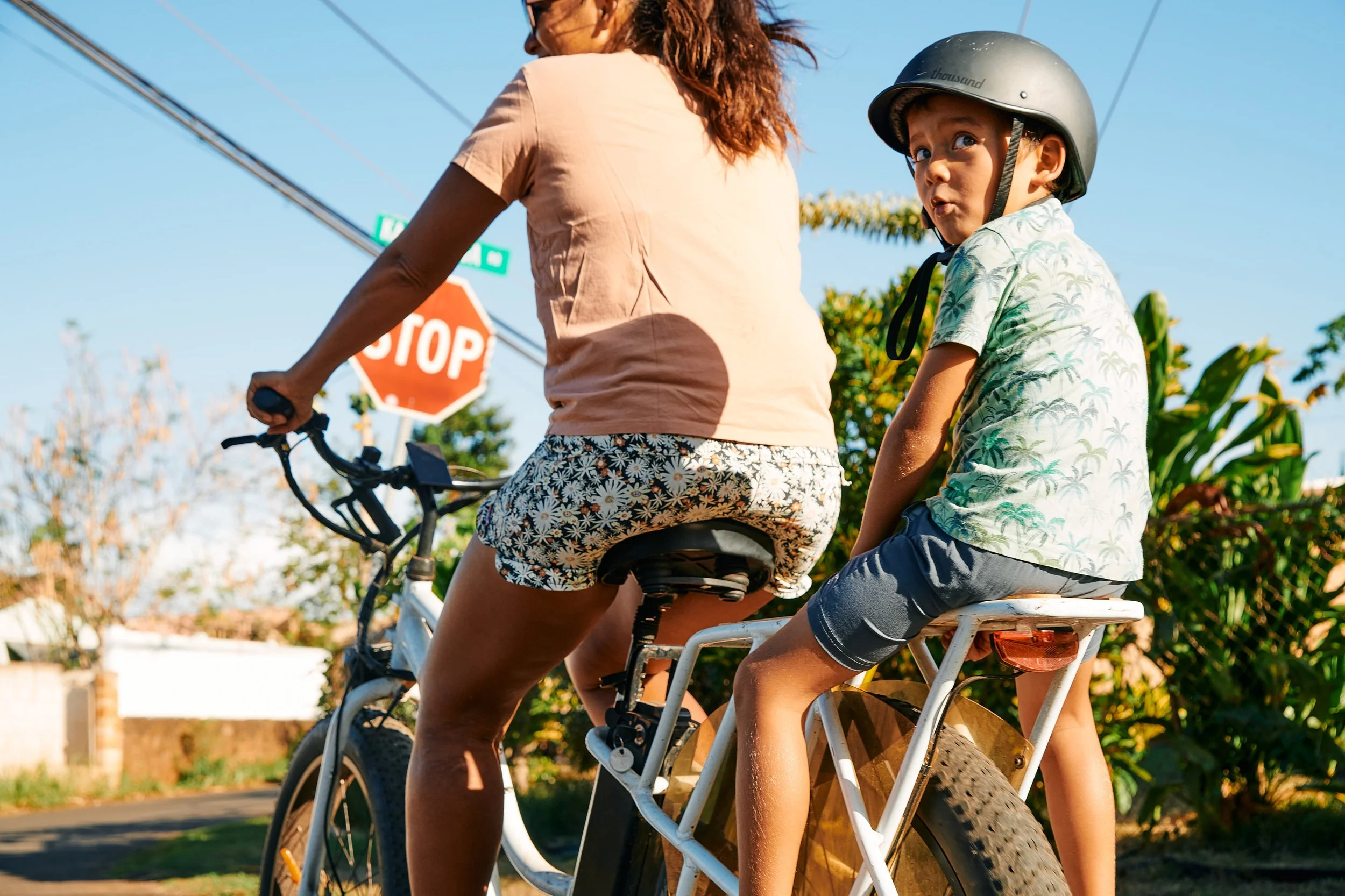 A woman and two children ride a bicycle together on a sunny day. The woman is pedaling, and a young girl is sitting on the back seat with her hands on her lap. The girl is wearing a helmet and casual clothes. They are passing a stop sign with trees a