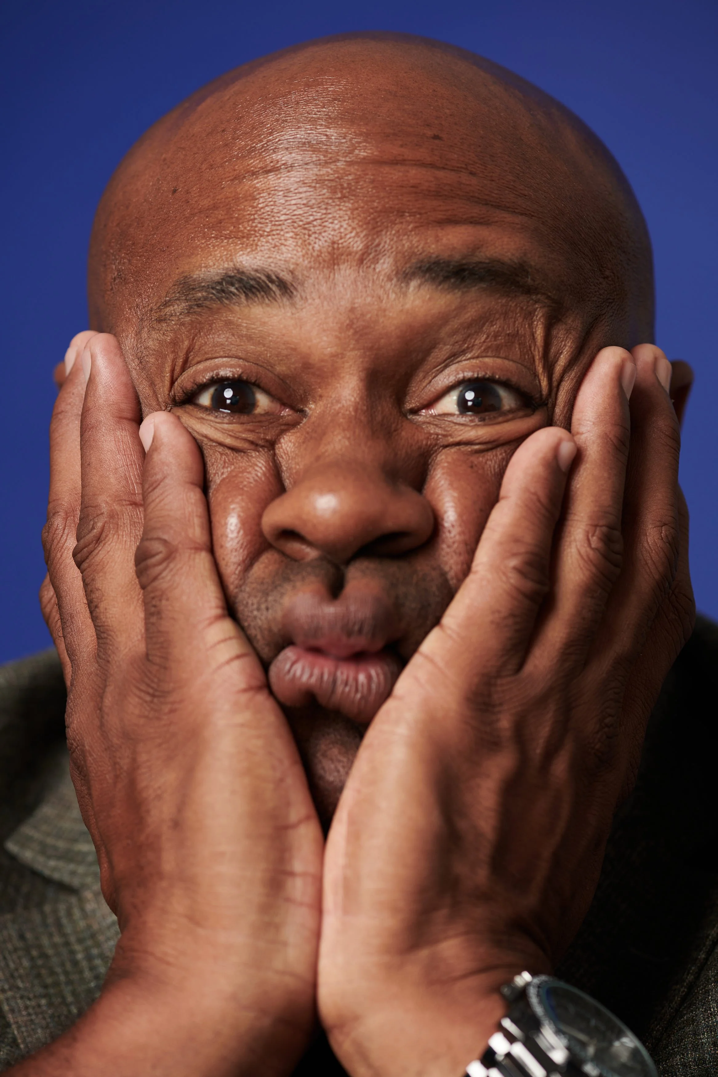 Close-up of a man making a surprised or playful face, pressing his cheeks with his hands, with wide eyes and puckered lips, against a blue background.