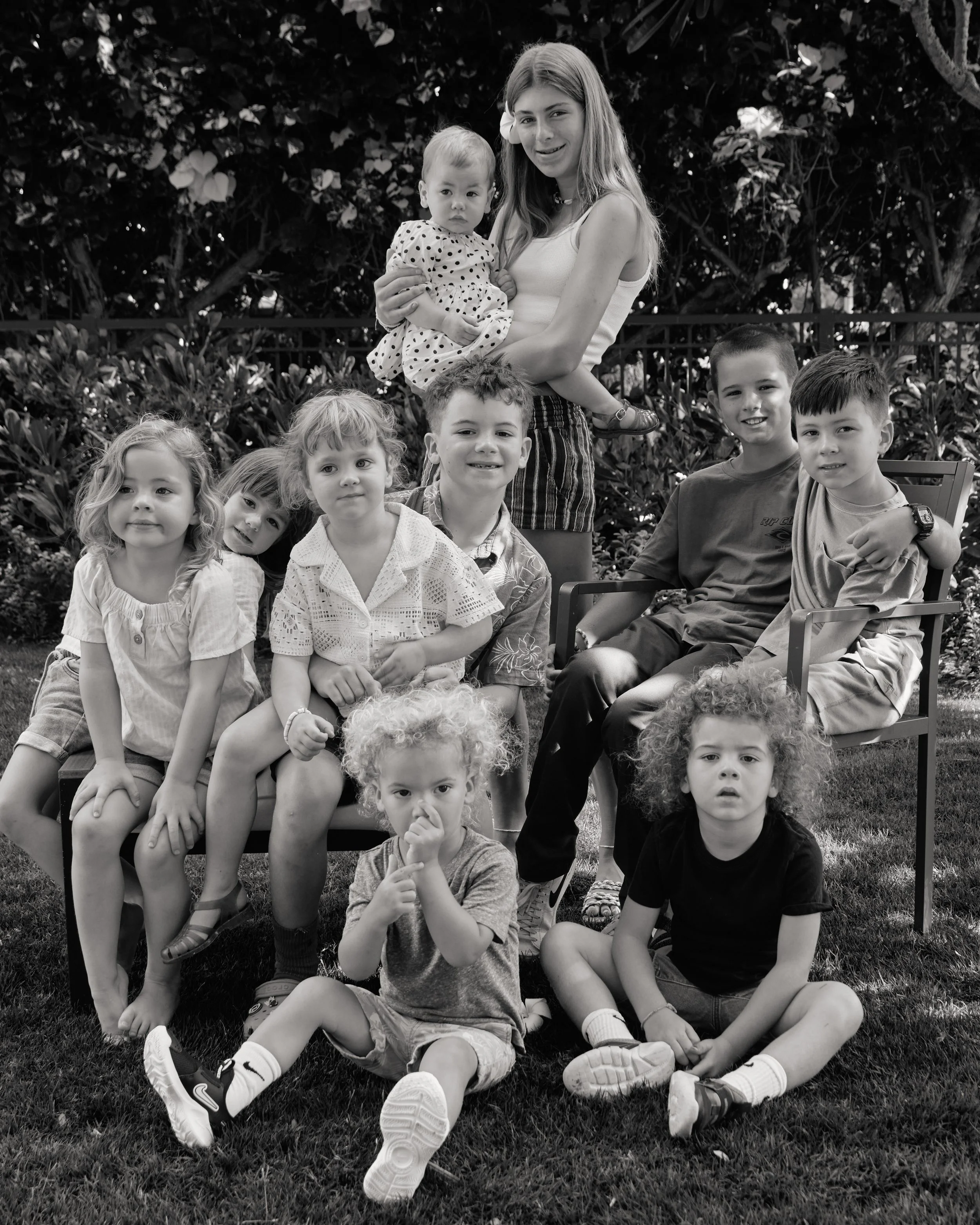 Group of children and a teenage girl outdoors, some sitting on chairs, others standing or sitting on the grass, in front of bushes and trees.