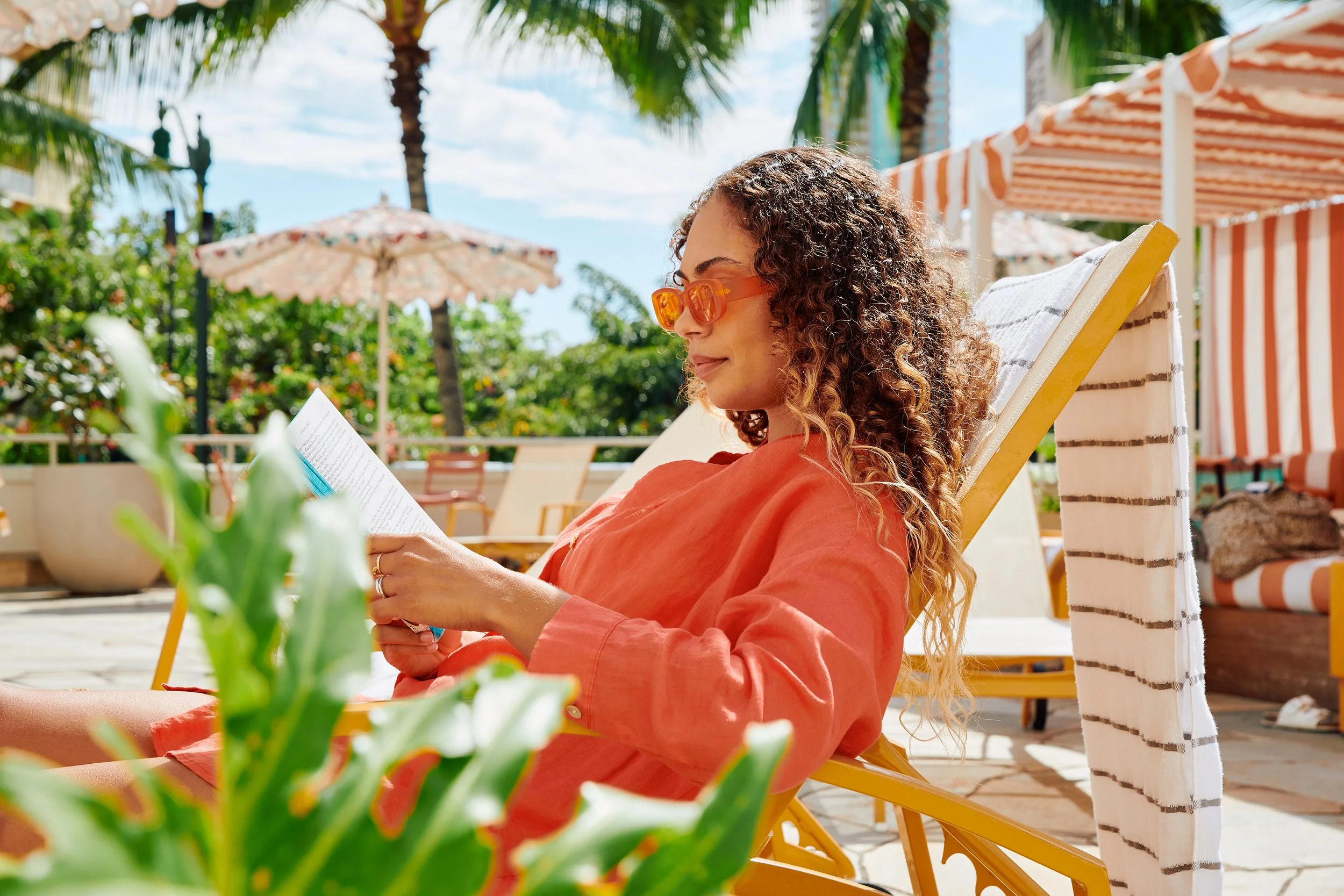 Woman with curly hair wearing orange sunglasses and a coral dress relaxing on a lounge chair at a poolside area, reading a book under a sunny sky with umbrellas and palm trees in the background.