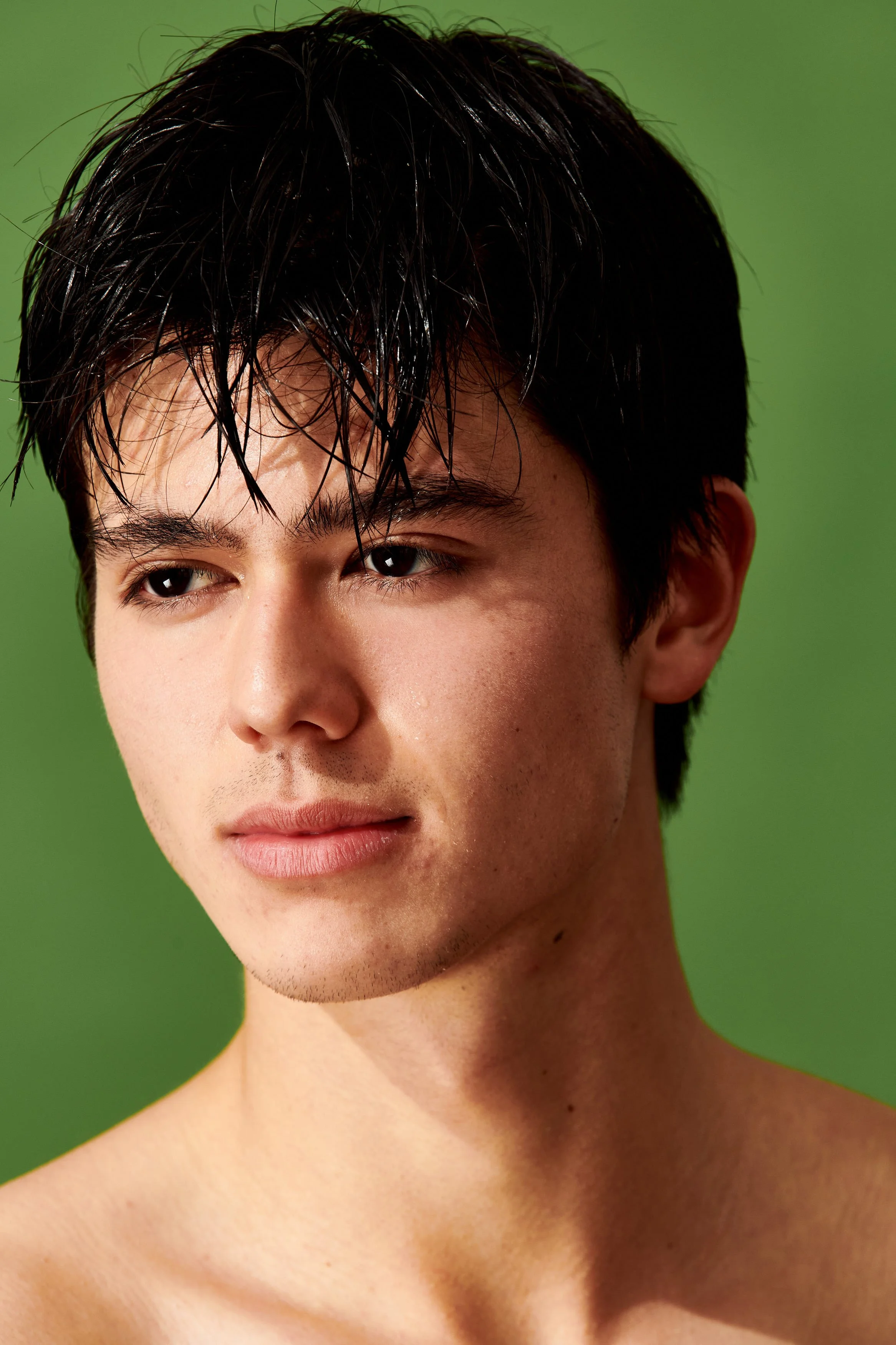 Close-up portrait of a young man with dark wet hair, lightly tanned skin, and a neutral expression, against a green background.