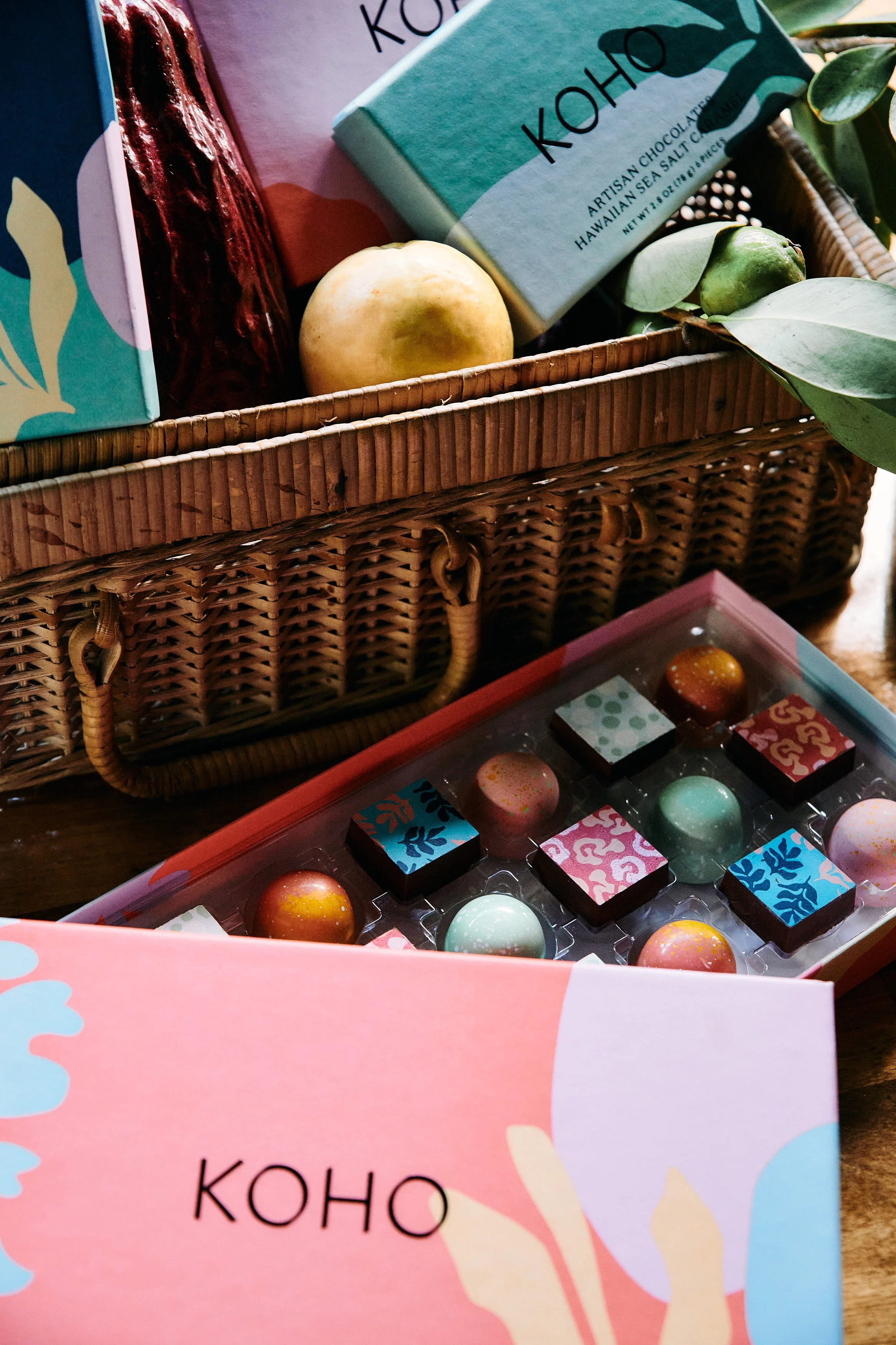A wicker basket containing boxes of artisan chocolates and two fruits, a yellow apple and a green guava. A box of chocolates with colorful, patterned wrappers and a box with the label 'KOHO' is also visible.