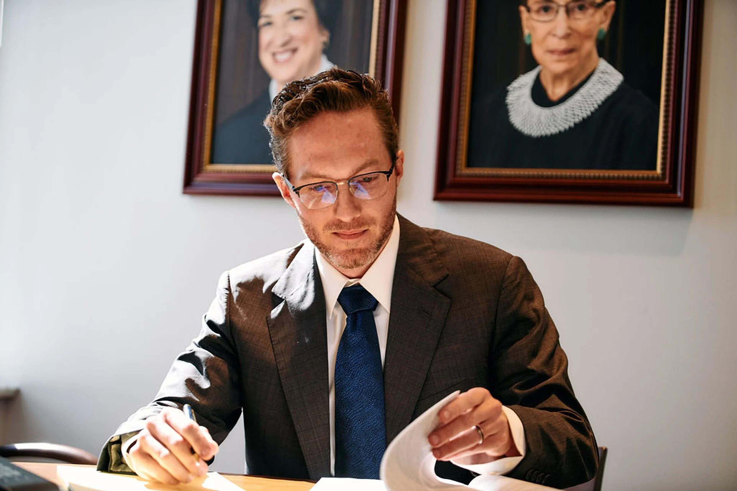 A man in a brown suit and blue tie sitting at a desk, reading a document with two portraits hanging on the wall behind him.