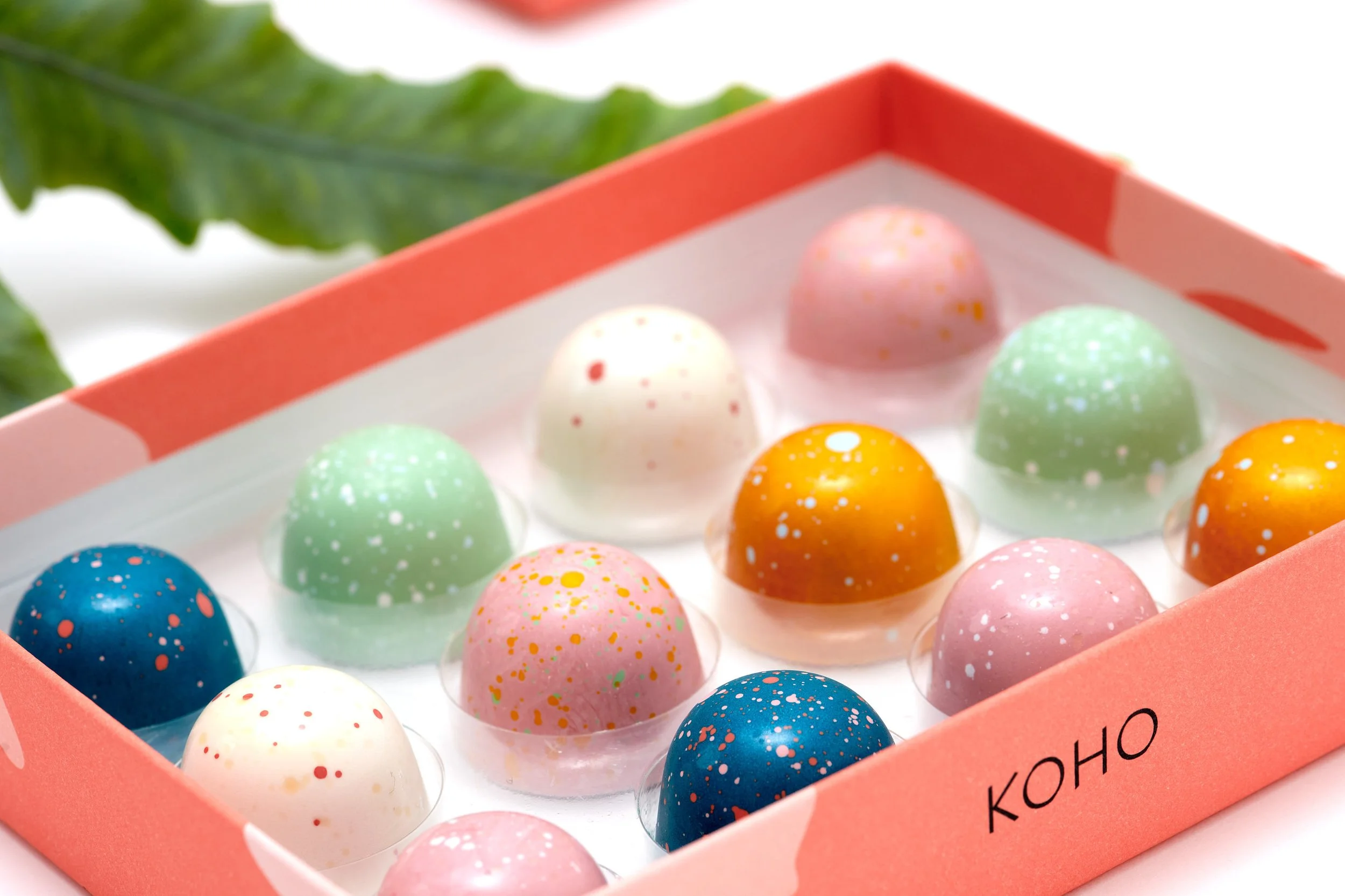 A pink box labeled 'KOHO' containing colorful, speckled mochi ice cream balls in various colors including blue, white, pink, green, yellow, and orange, with a leafy background.