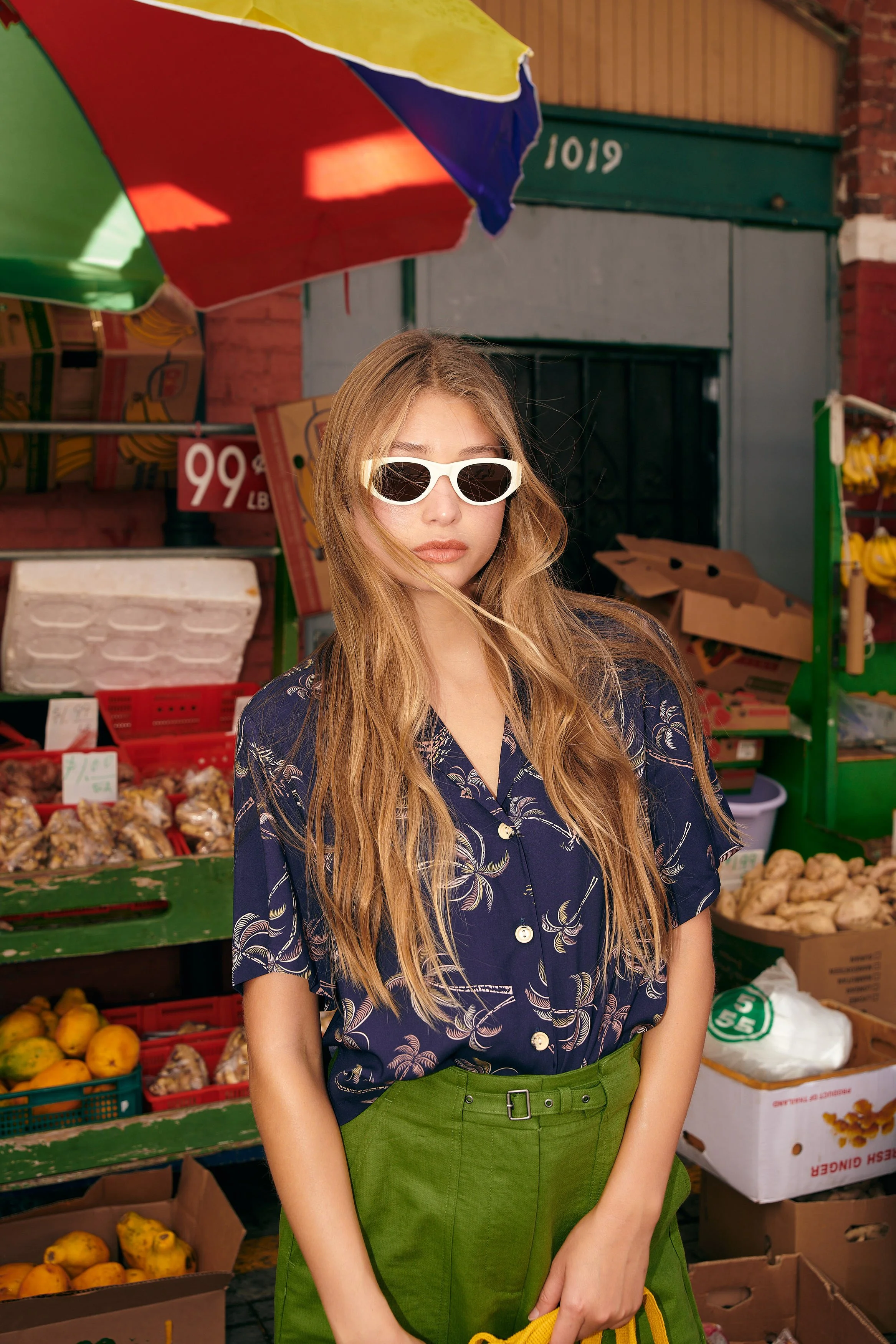A young woman with long blonde hair wearing white sunglasses, a navy blue shirt with a floral pattern, and green pants stands at an outdoor market stall with fruits and vegetables.