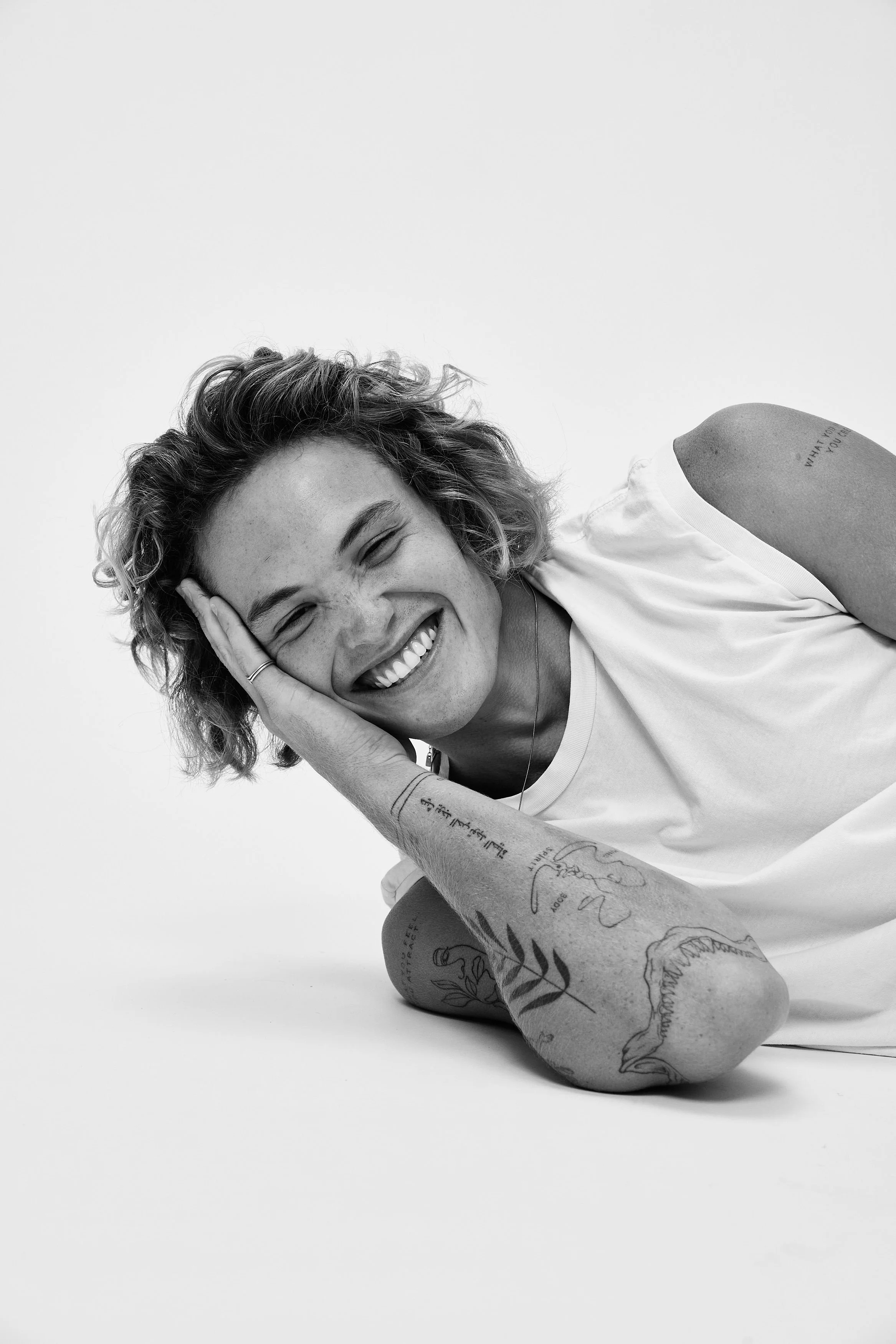 A young woman lies on her side, smiling, with her head resting on her hand, wearing a white T-shirt, with visible tattoos on her arm and neck, in a studio with a plain background.