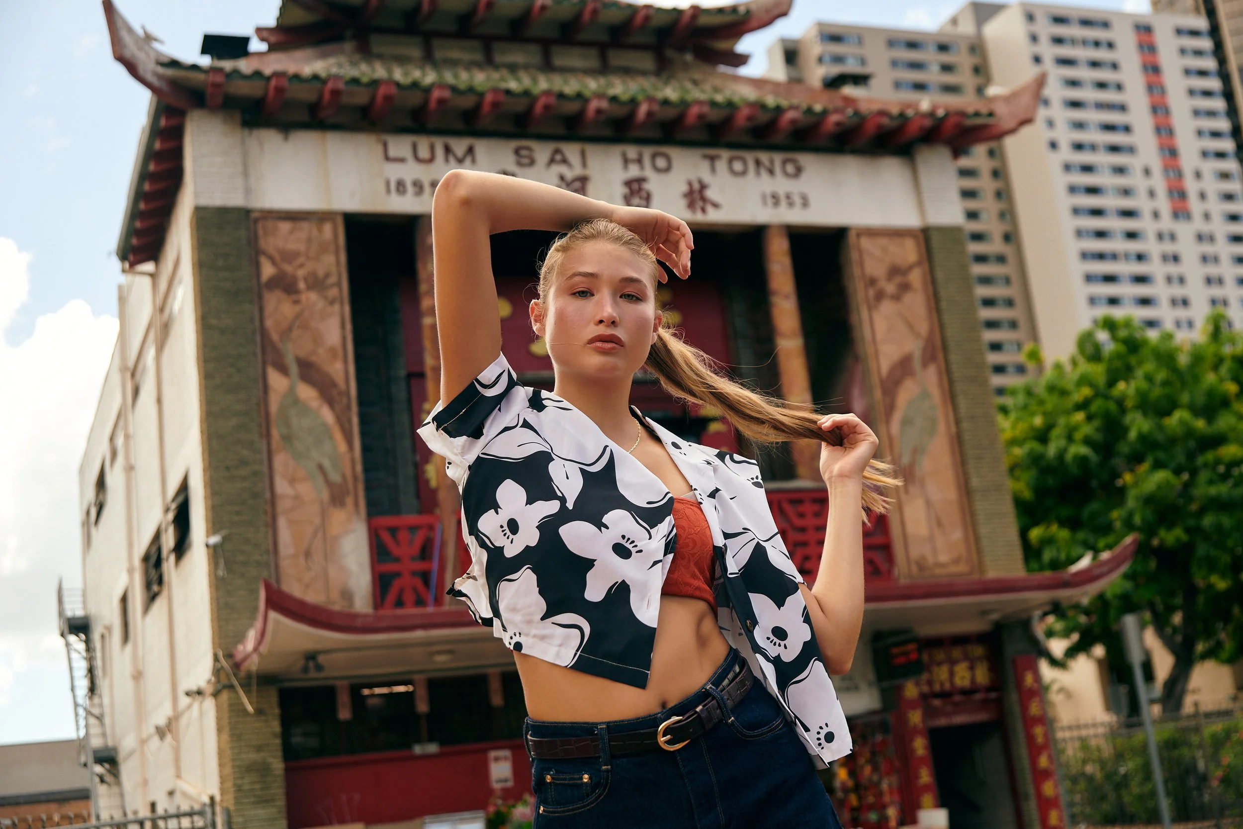 Young woman with long hair posing in front of a traditional Chinese-style building, wearing a black and white floral shirt and a red top underneath.