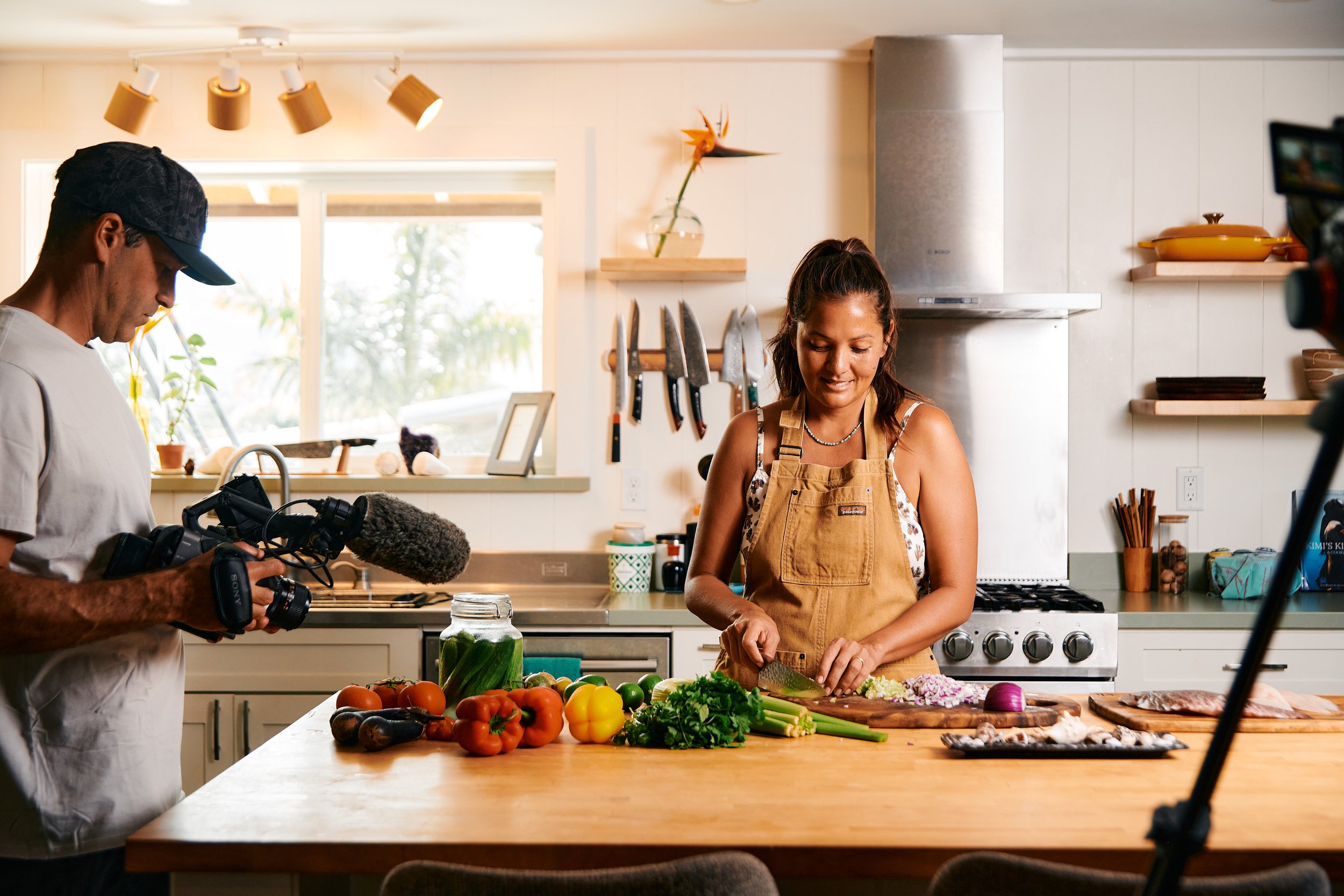 A woman in a beige apron chopping vegetables while being filmed by a man holding a camera with a microphone in a kitchen.
