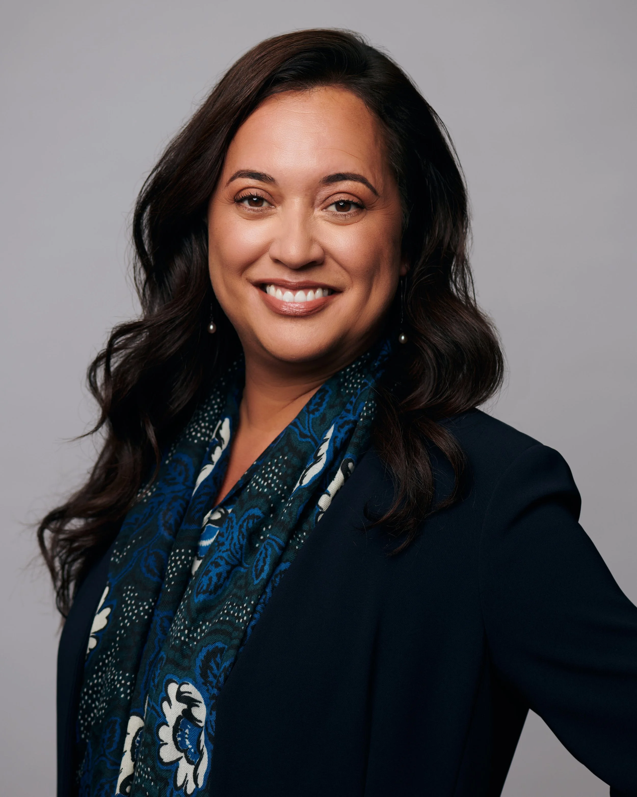A woman with long dark hair, smiling, wearing a navy blazer and a blue patterned scarf, against a plain light gray background.