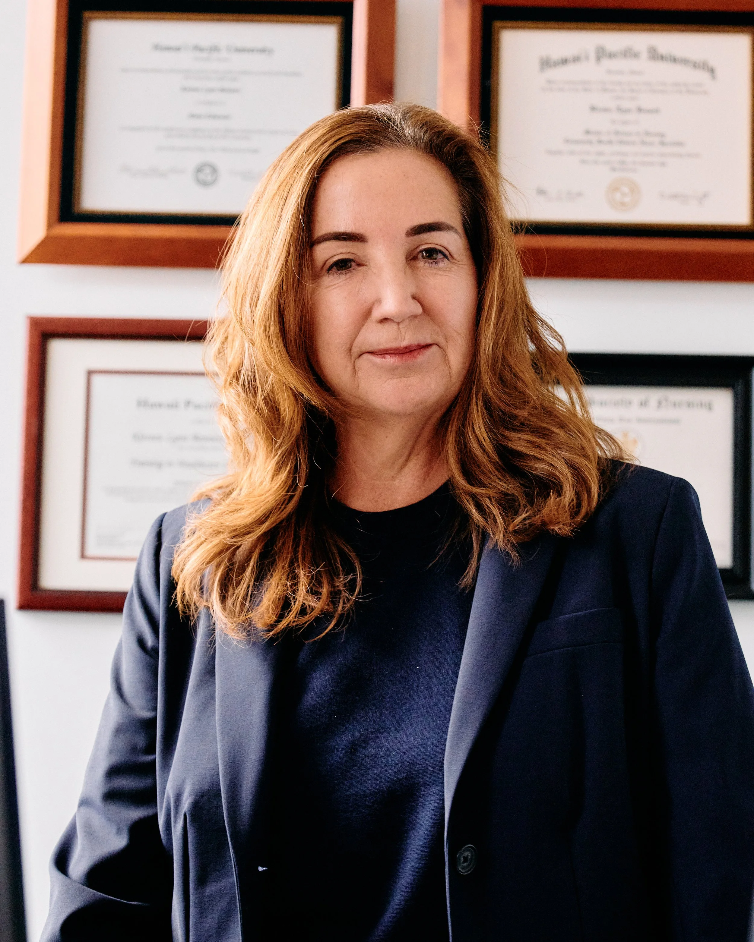 A woman with long, wavy auburn hair and fair skin, wearing a navy blazer and a black top, standing in front of a wall with framed diplomas.