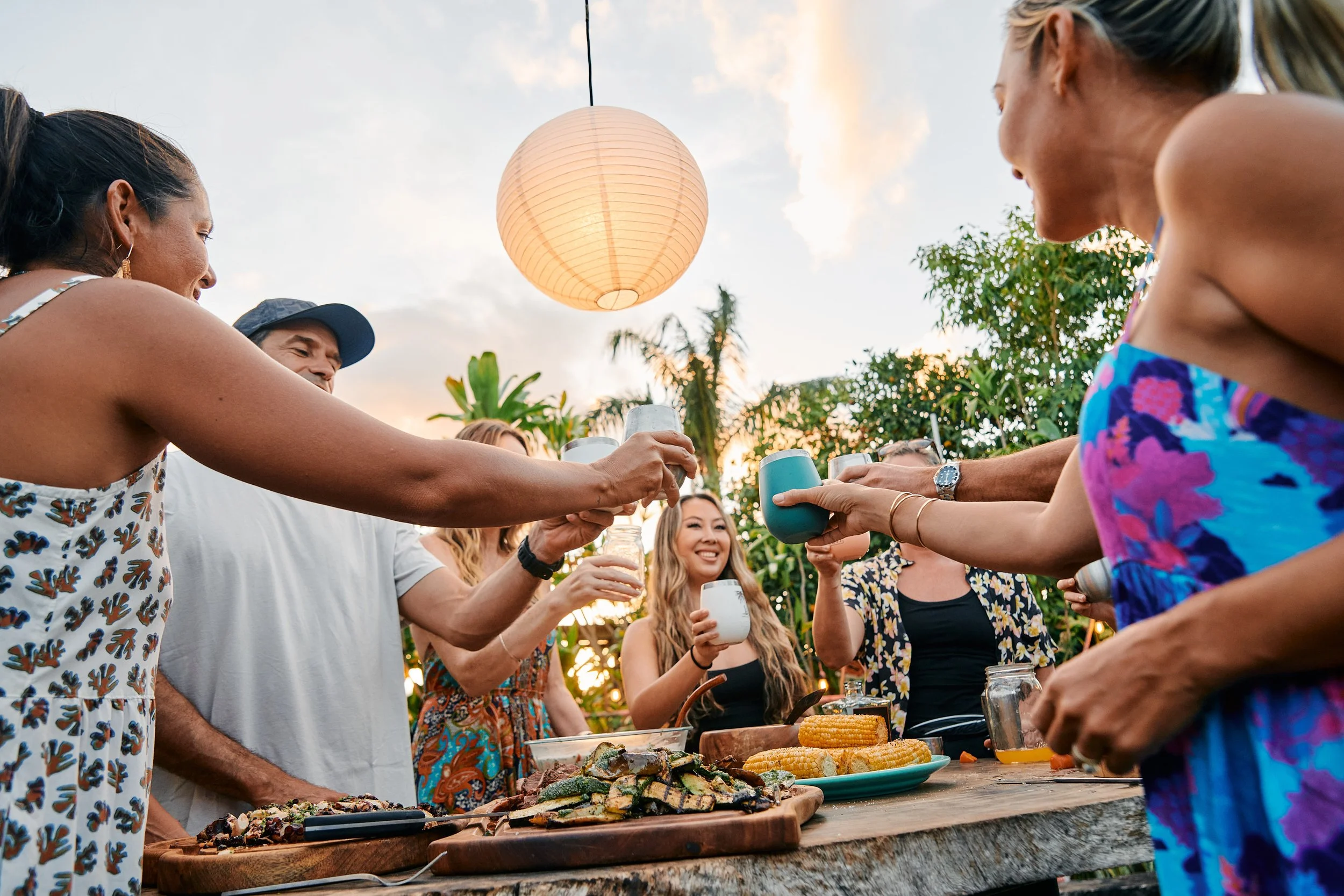 People gathered outdoors celebrating with drinks and food, raising glasses in a toast during sunset.