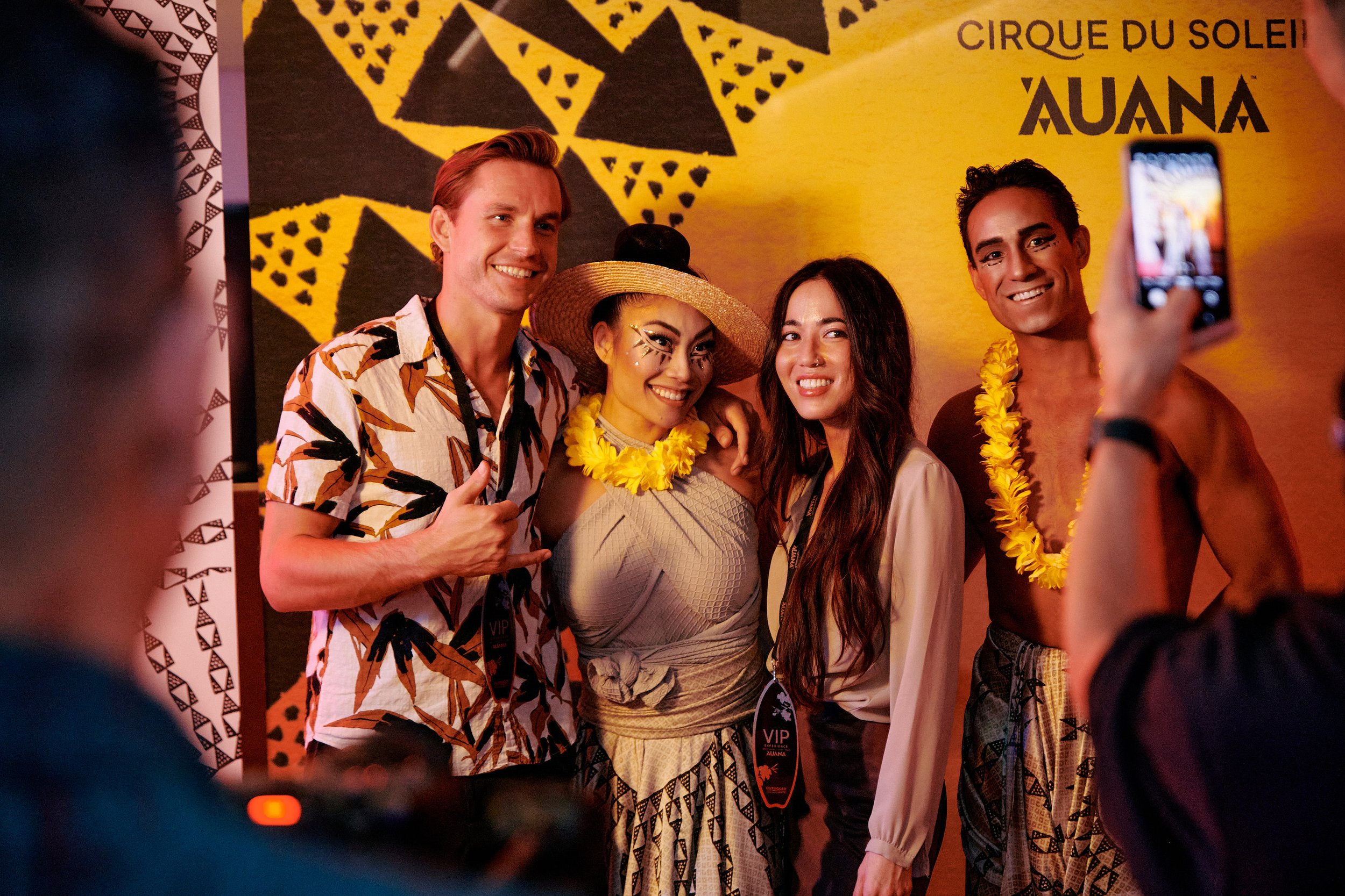 Four people posing for a photo at a Cirque du Soleil event, two men and two women, with tropical-themed attire and accessories, standing in front of a yellow backdrop with black patterns.