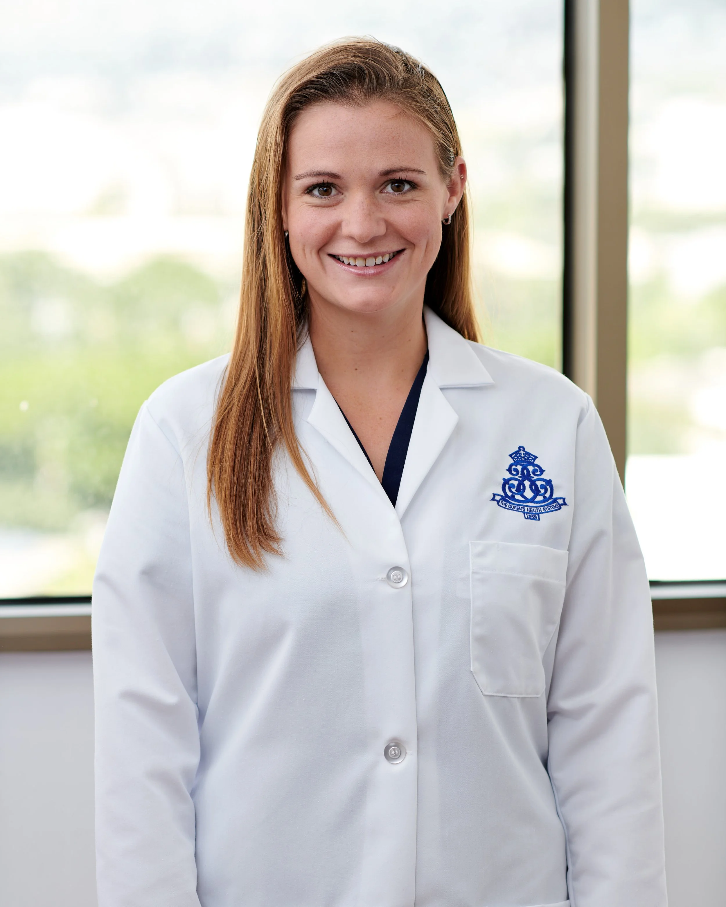 A woman wearing a white lab coat with a blue embroidered logo, standing indoors in front of large windows with a view of greenery outside, smiling at the camera.