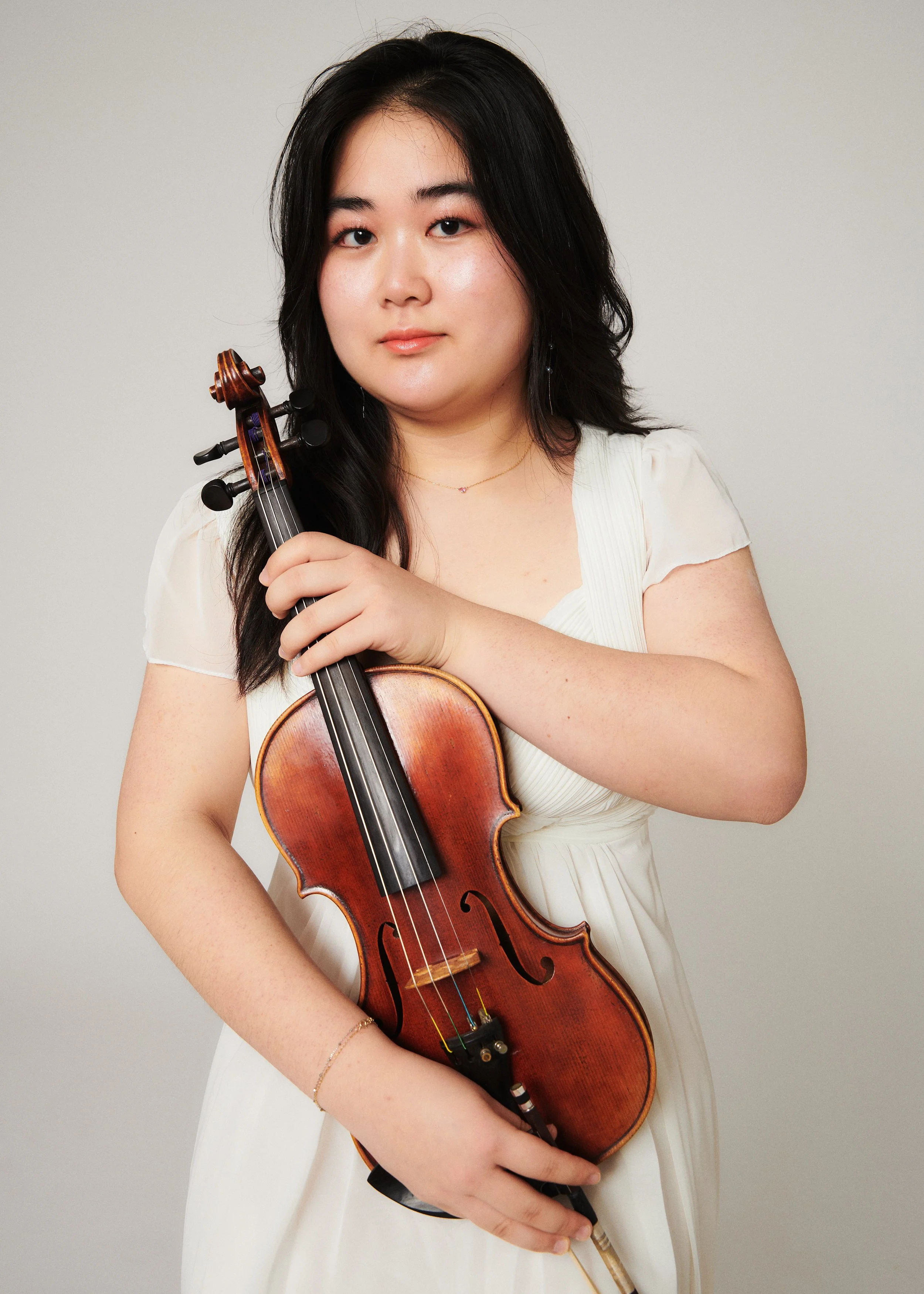 A woman with long dark hair holding a violin in front of a plain gray background.