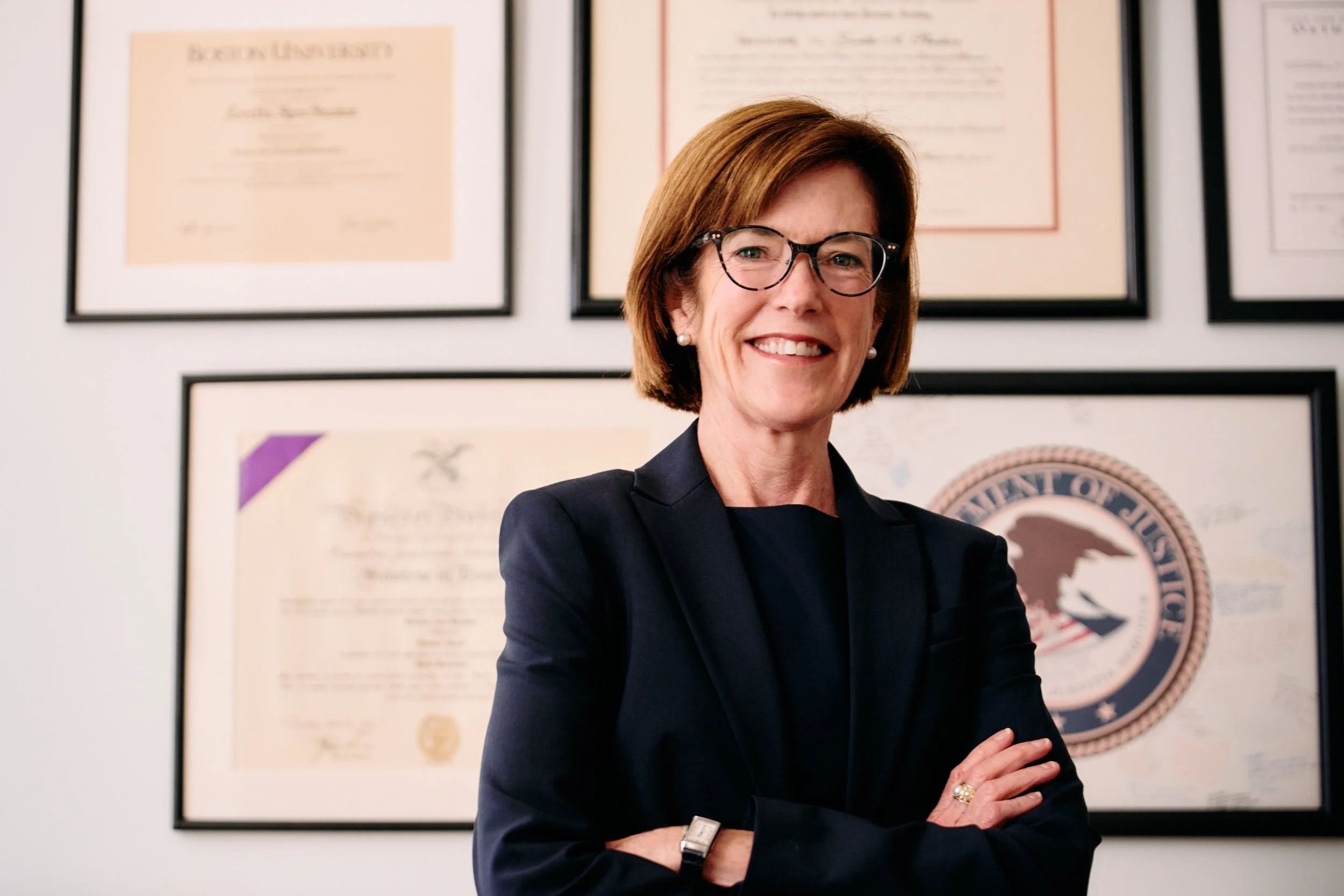 A woman with glasses, wearing a black blazer, stands with her arms crossed in front of a wall with framed diplomas and certificates, including a United States Department of Justice seal.