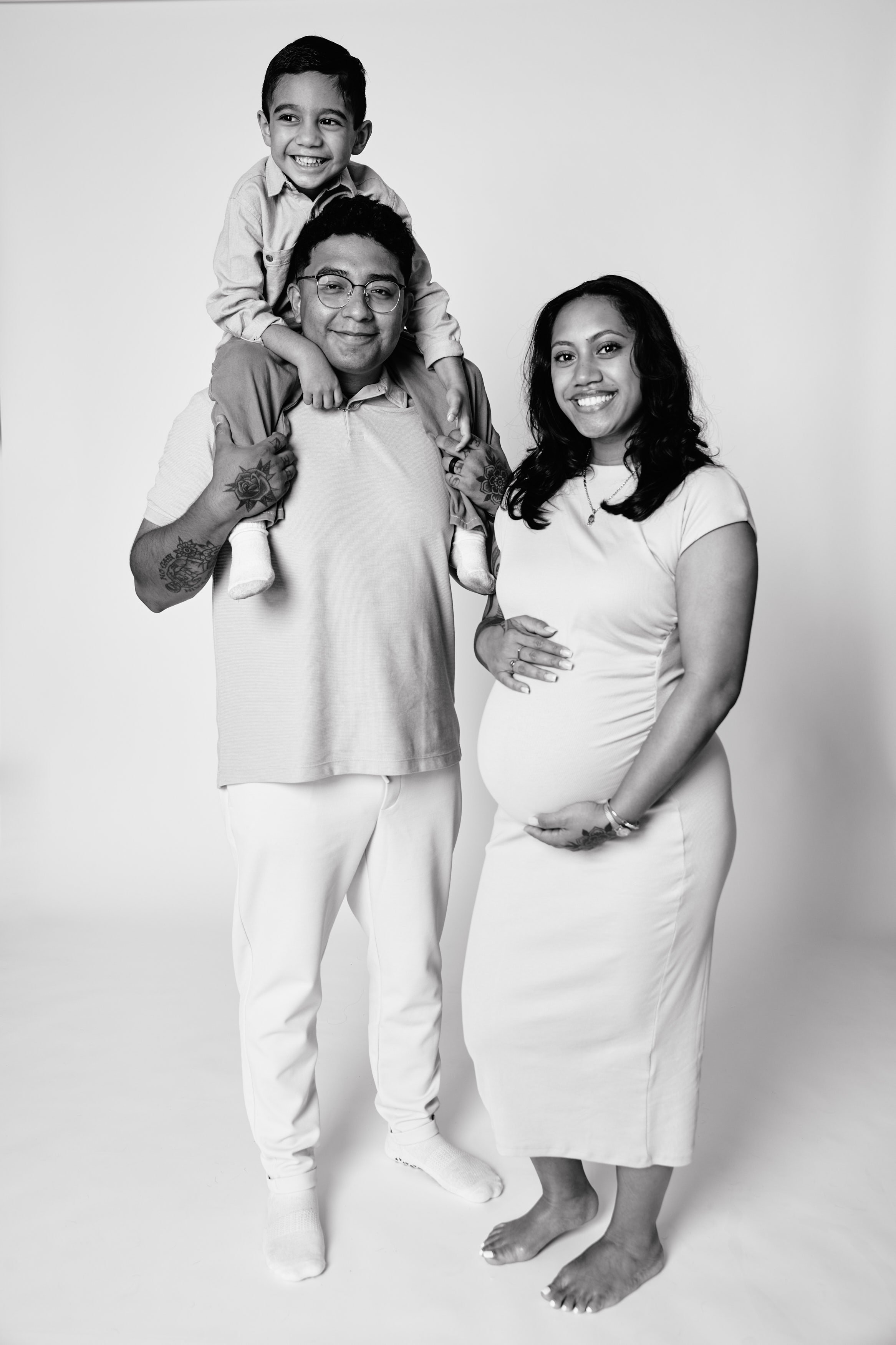 Family portrait of an expectant mother, father, and their young son, all smiling, standing against a plain background.