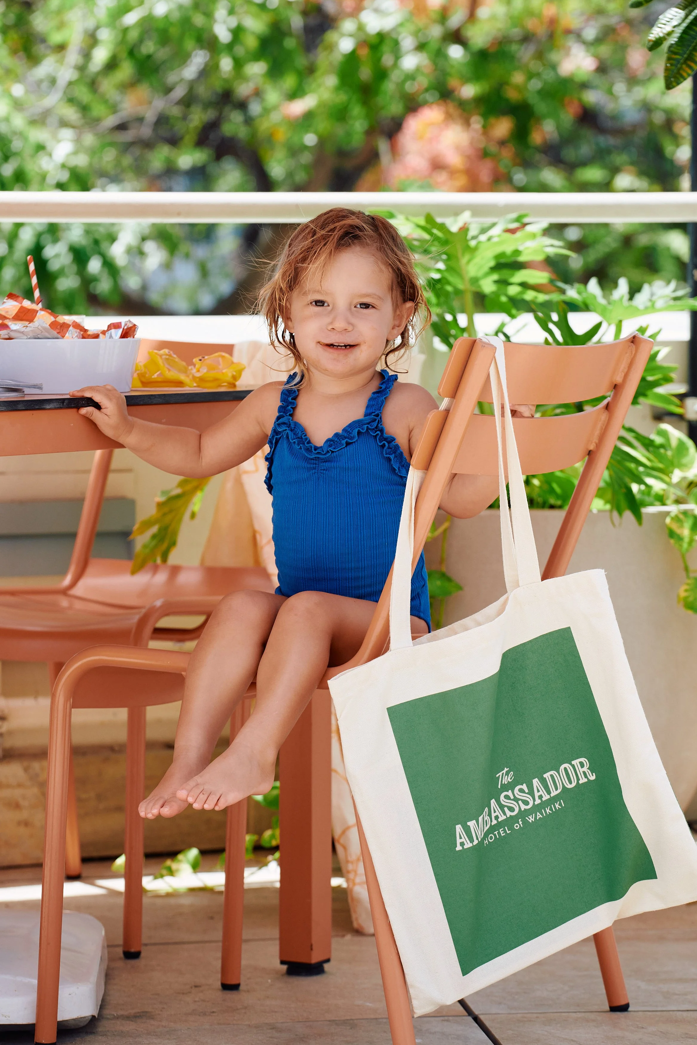 A young girl with red hair in a blue swimsuit sitting on a peach-colored chair on a balcony, holding onto the back of the chair, with a tote bag hanging from the chair's side. The balcony has green plants in the background, and a table with snacks an