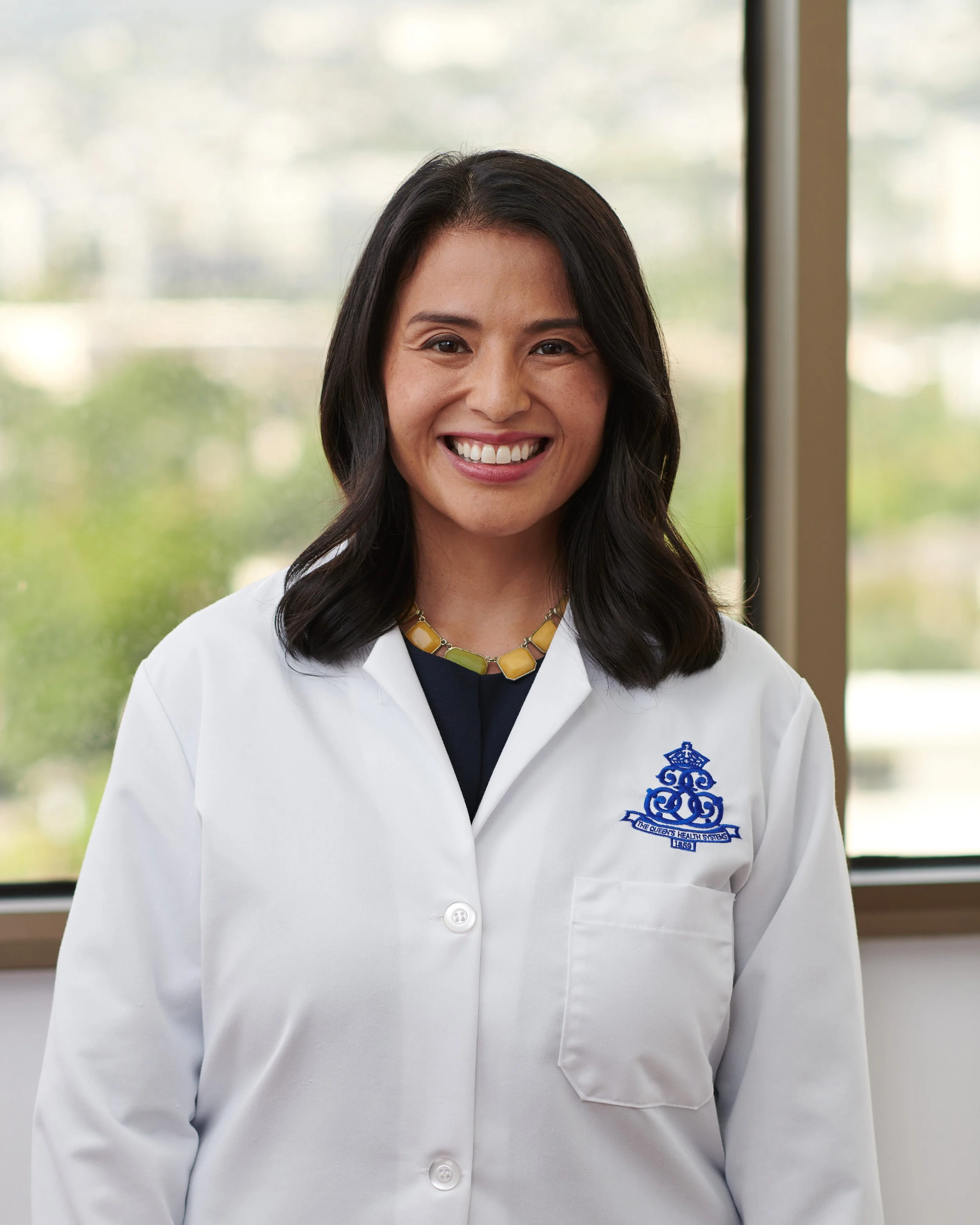 A woman smiling in a white medical coat with a blue emblem, standing indoors near a window with a blurred outdoor background.