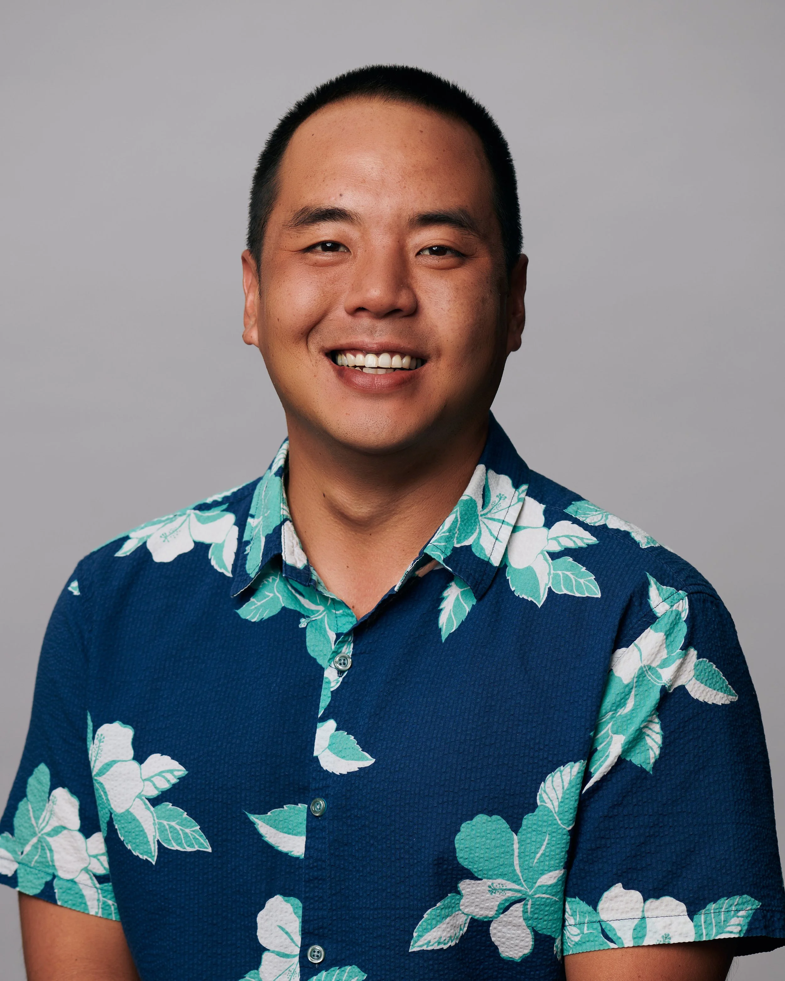 A smiling man with short black hair wearing a blue Hawaiian shirt with white and green floral patterns, against a plain gray background.