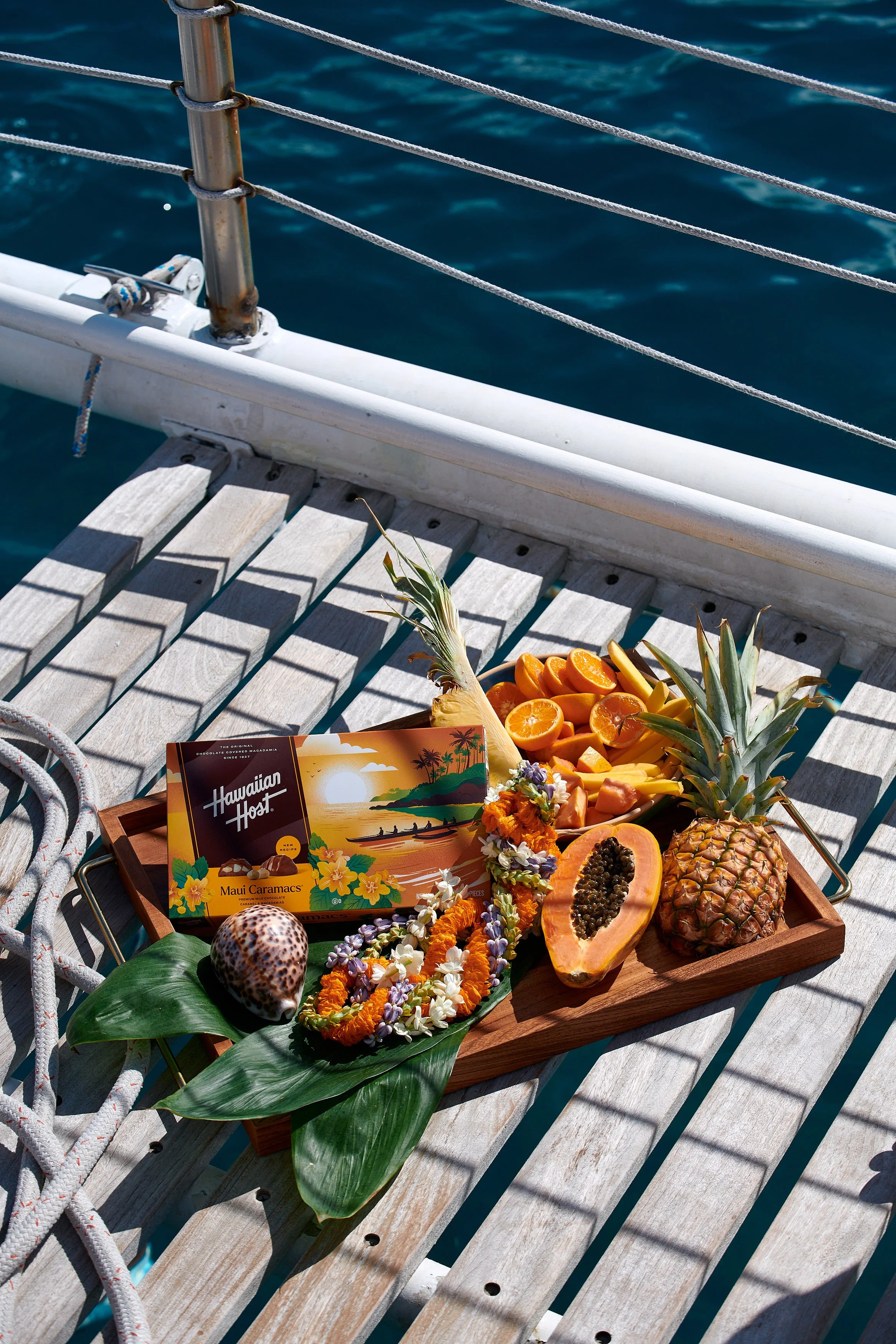 A wooden tray of tropical fruits, including pineapple, papaya, orange slices, and a banana, along with a Hawaiian Host Maui Caramacs chocolate box, marigold and other flower leis, and a seashell, placed on a deck with a boat railing and water in the 
