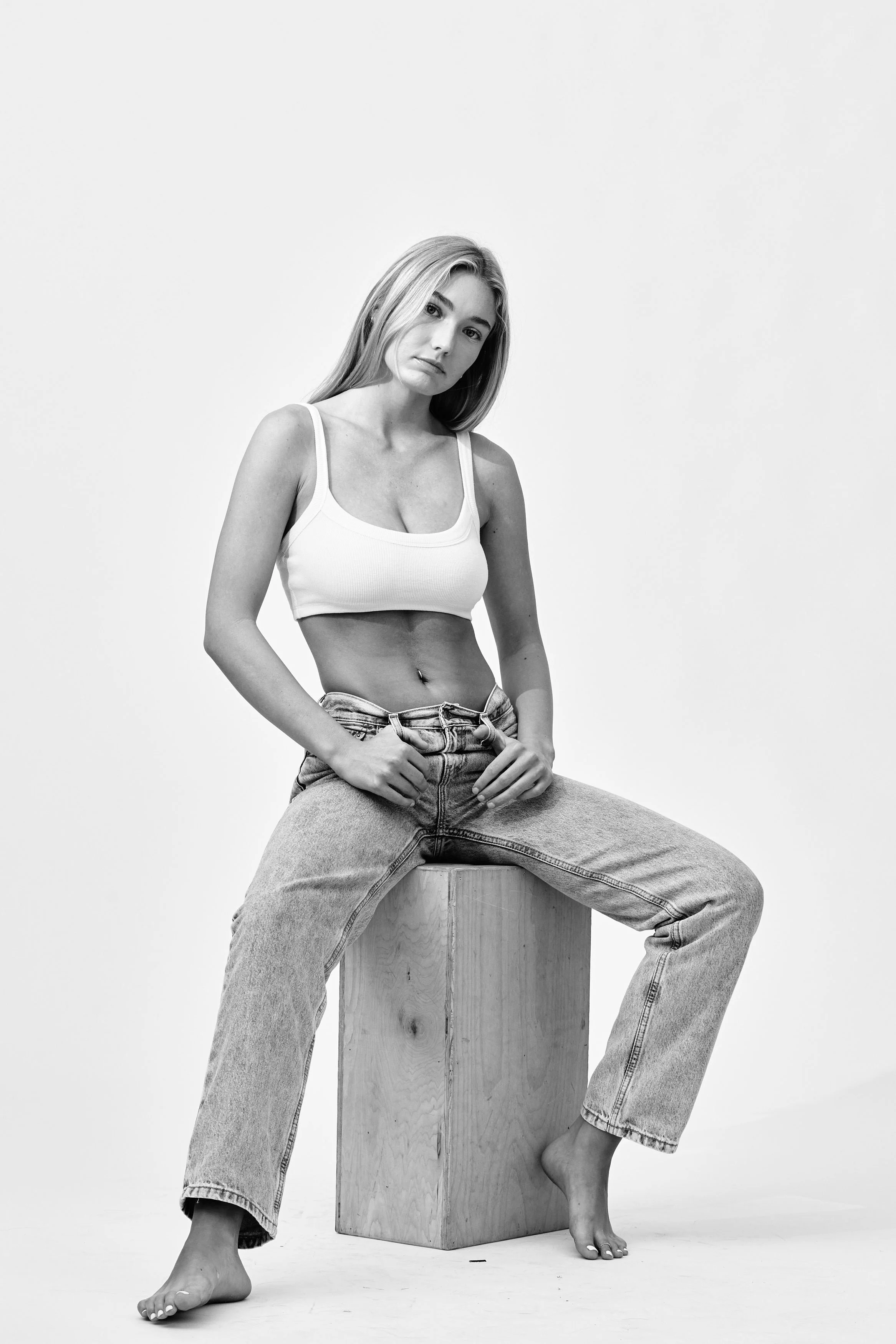 Black and white photo of a young woman sitting on a wooden cube, wearing a white crop top and loose jeans, with a neutral expression, barefoot, against a plain background.