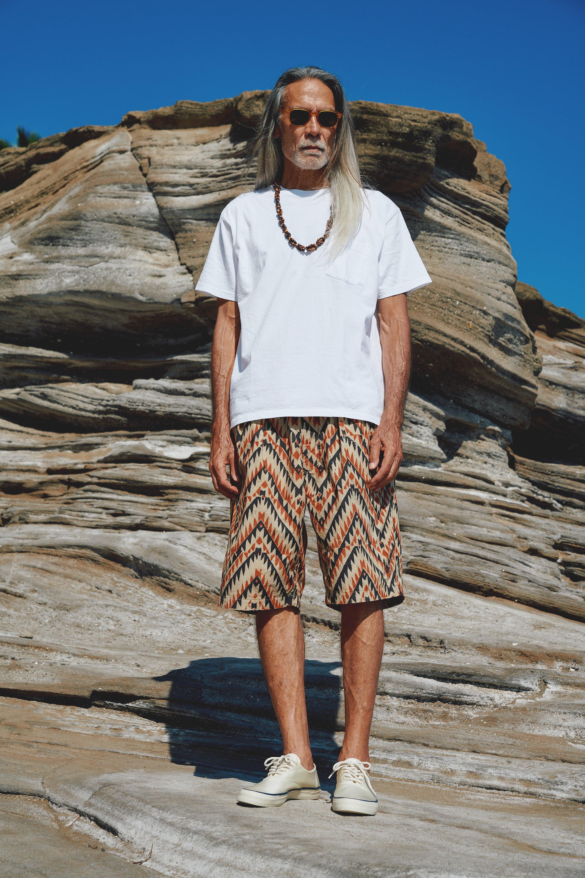 An elderly man with long gray hair wearing sunglasses, a white t-shirt, patterned shorts, and beige shoes standing on a rocky landscape with a clear blue sky in the background.