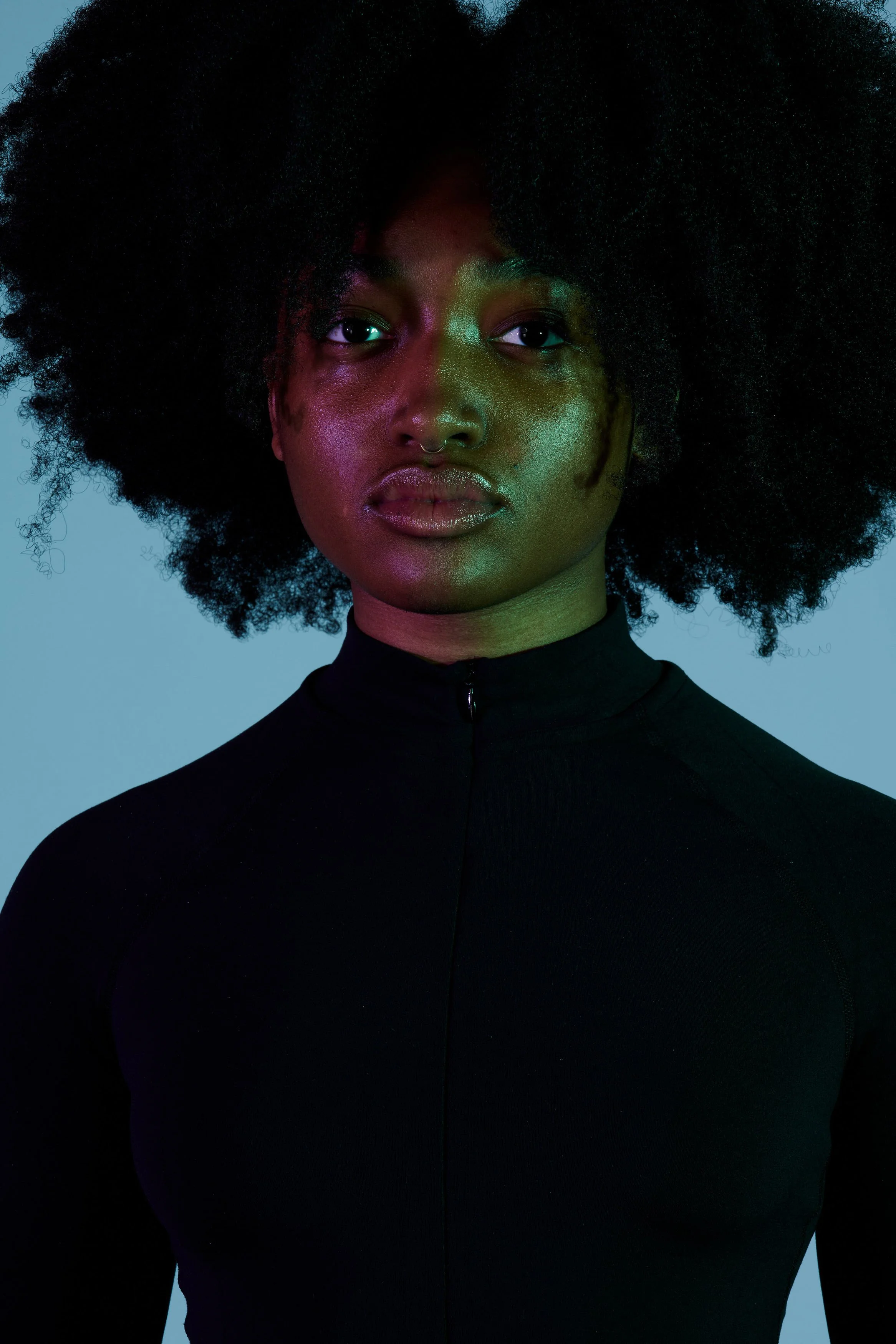Portrait of a Black woman with natural curly hair, wearing a black zip-up top, with colorful lighting on her face against a gray background.
